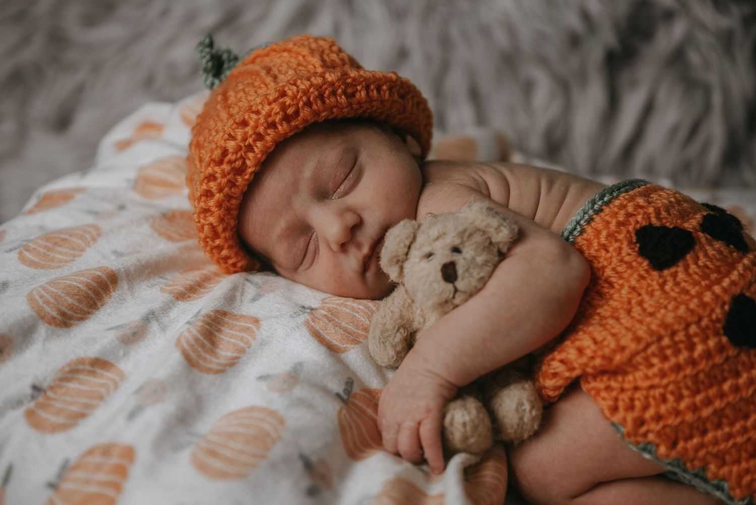 Sleeping baby wearing orange knit hat cuddling with teddy bear on white blanket.