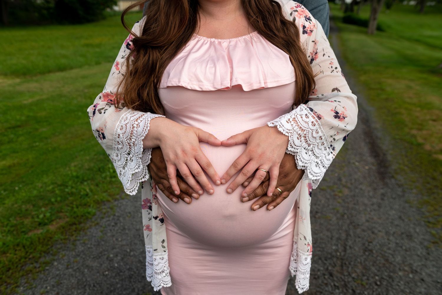 Maternity portrait session showing a pink dress and white lace overlay in a green field.