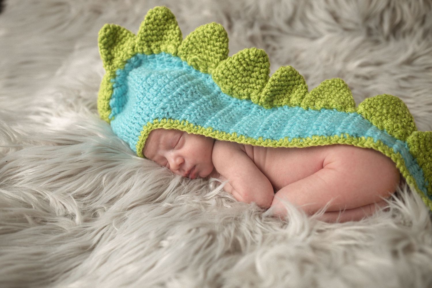 Baby in green and blue dinosaur costume resting on plush grey surface.
