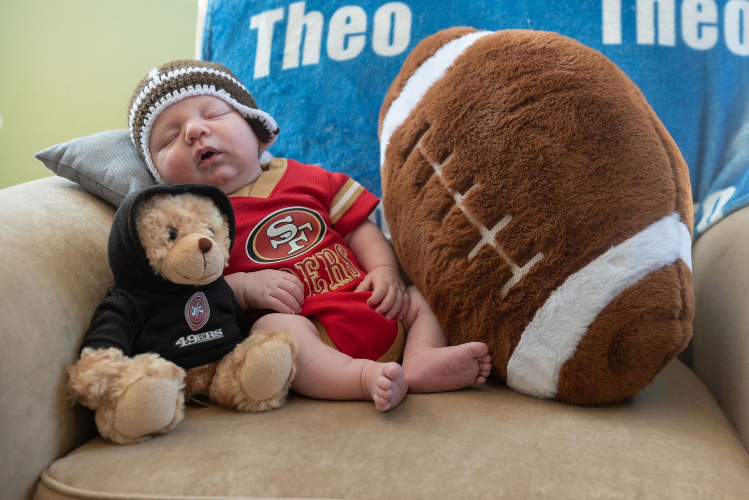 Baby resting on couch with football and teddy bear plush toys.