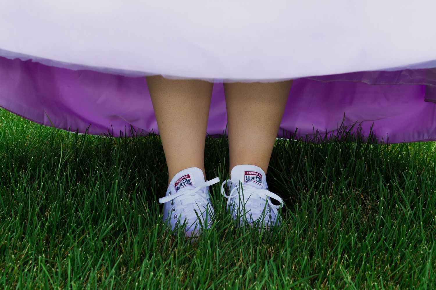 White shoes peek out from under a purple dress on green grass.