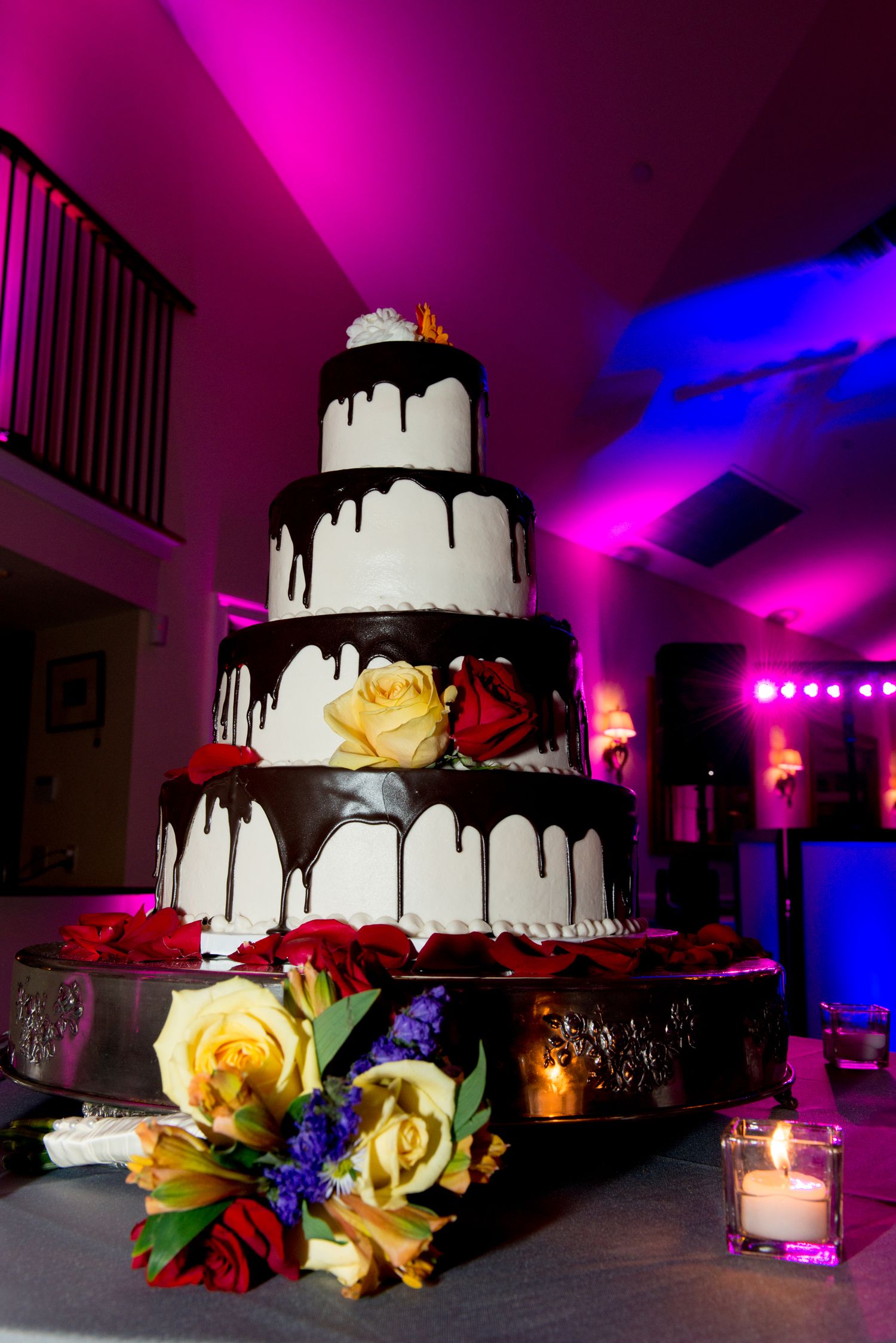 Elegant white wedding cake with black drip details and colorful floral decorations under dramatic purple lighting.