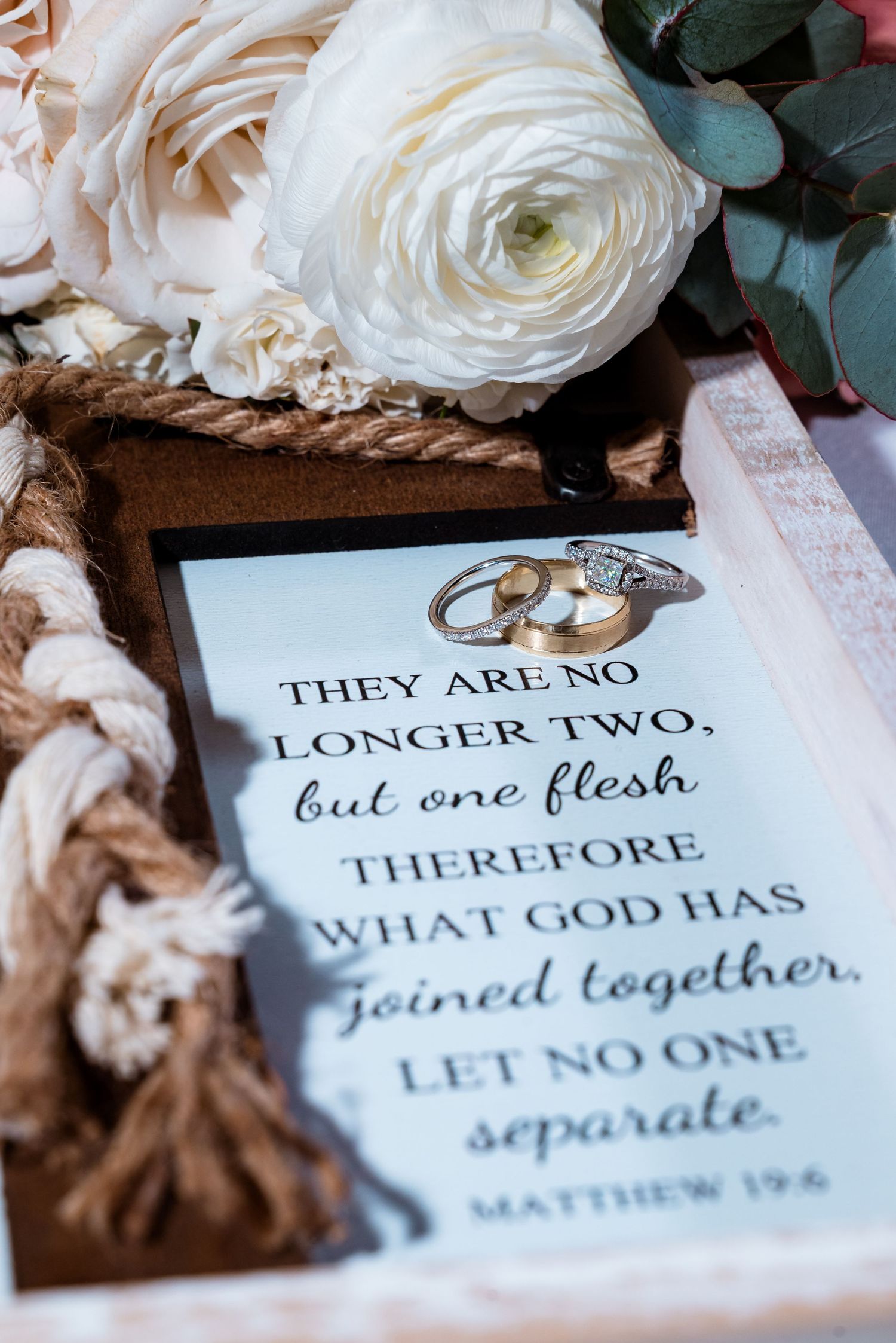 Wedding rings and white roses arranged with biblical scripture from Matthew 19:6.