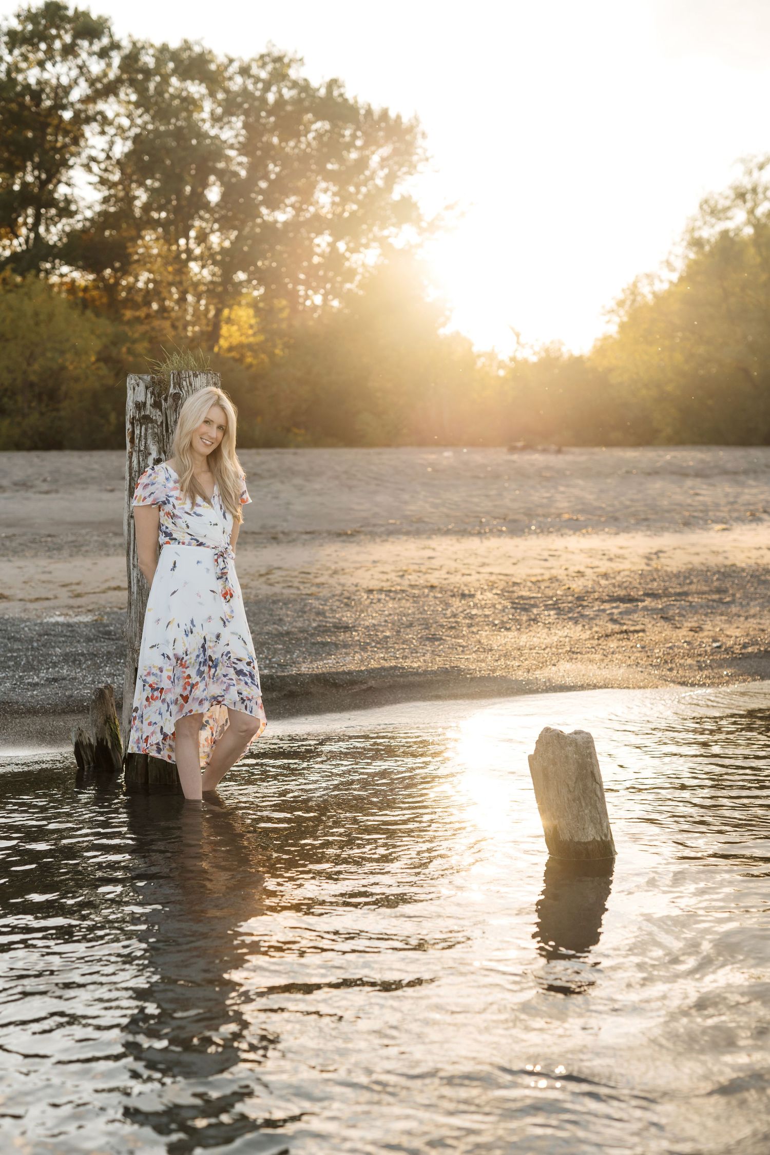 Michelle Hugill standing in shallow lake water at sunset, wearing a white floral dress
