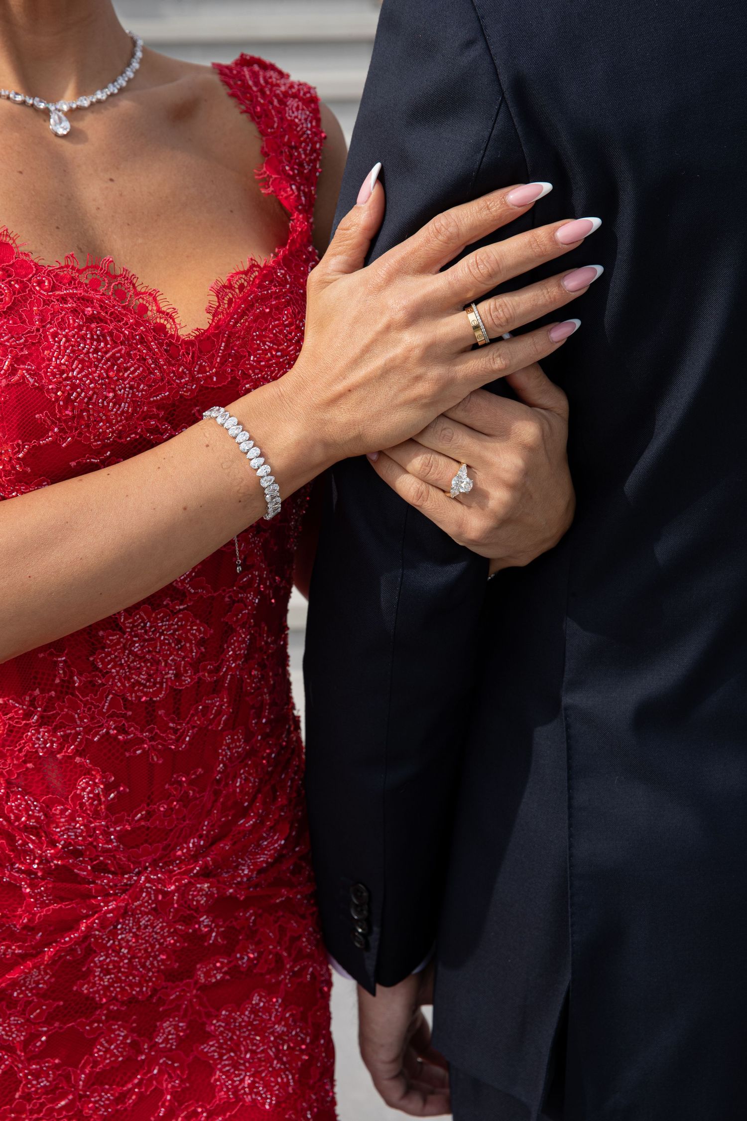 Woman in red lace dress with diamond ring and bracelet holds man's arm in dark suit.