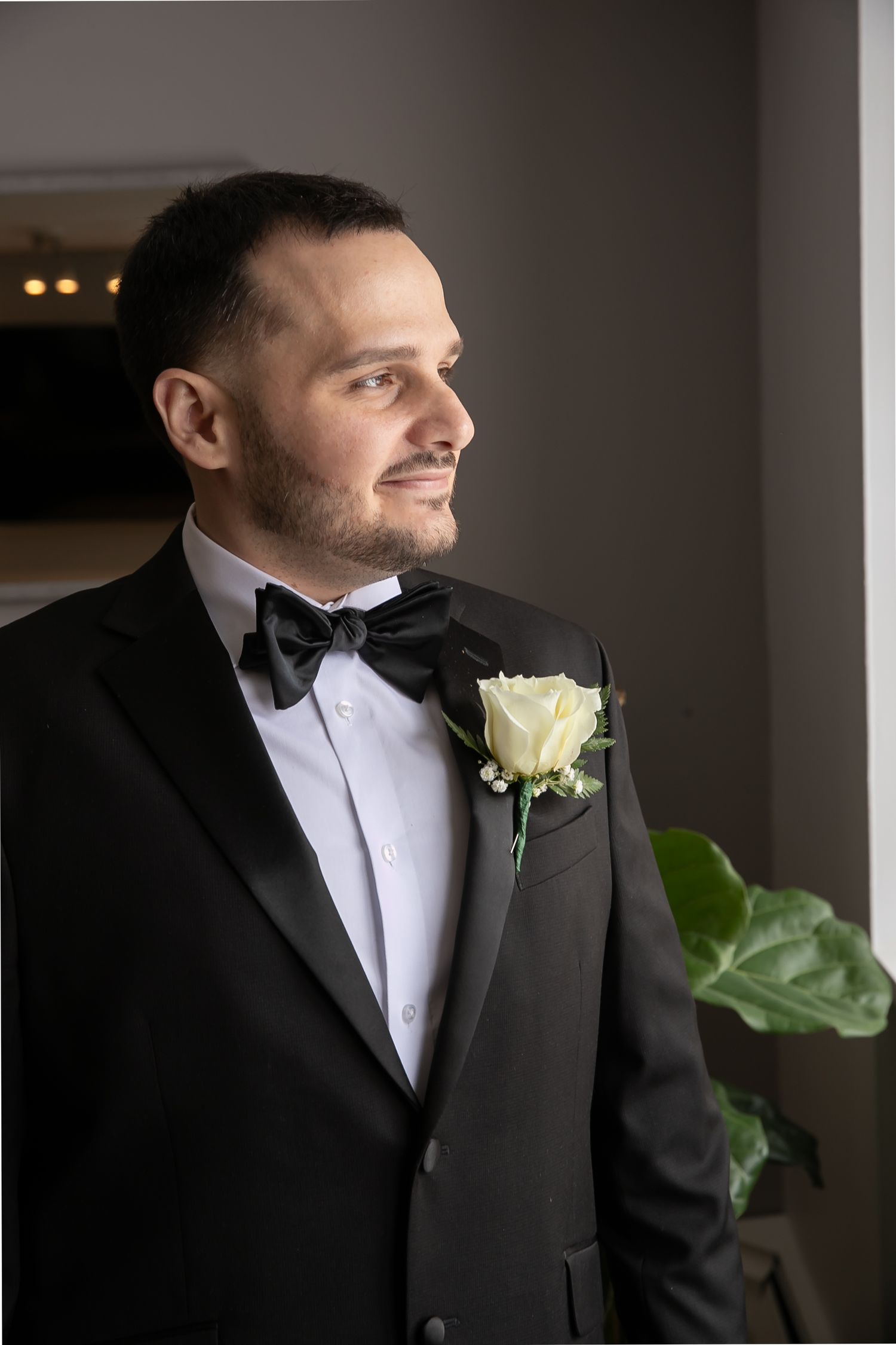 Groom in black tuxedo with white rose boutonniere gazing thoughtfully toward window on wedding day.