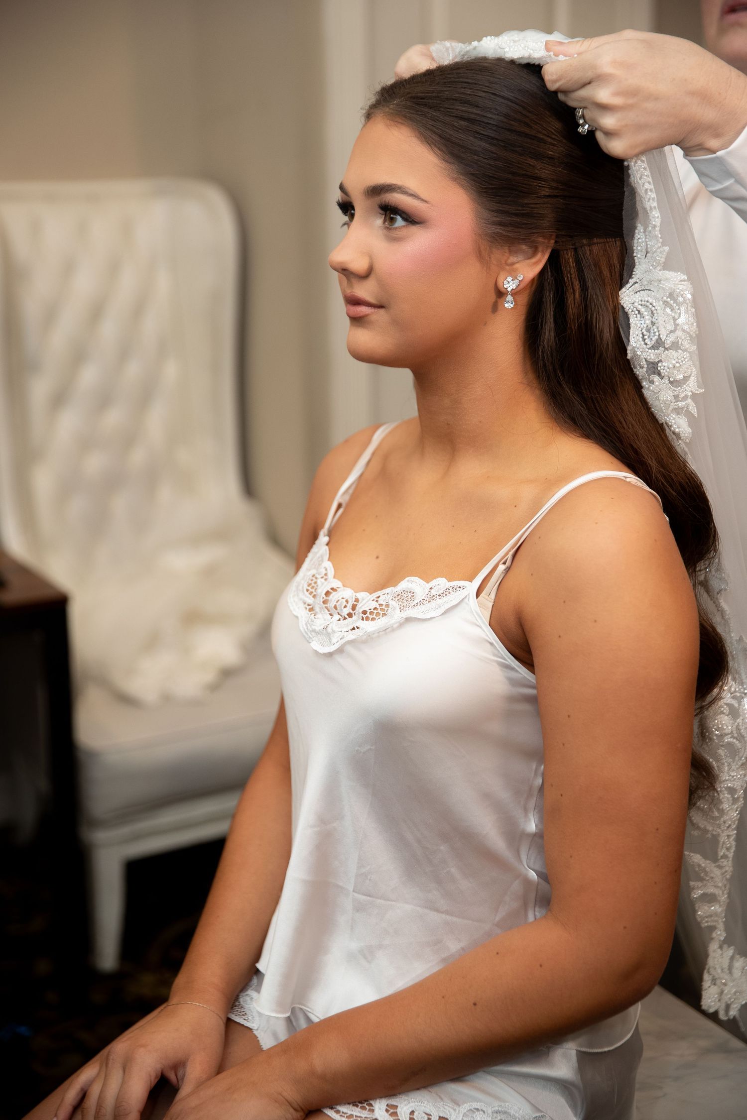 Bride in white satin slip dress getting veil placed by stylist, with elegant hair and pearl earrings.