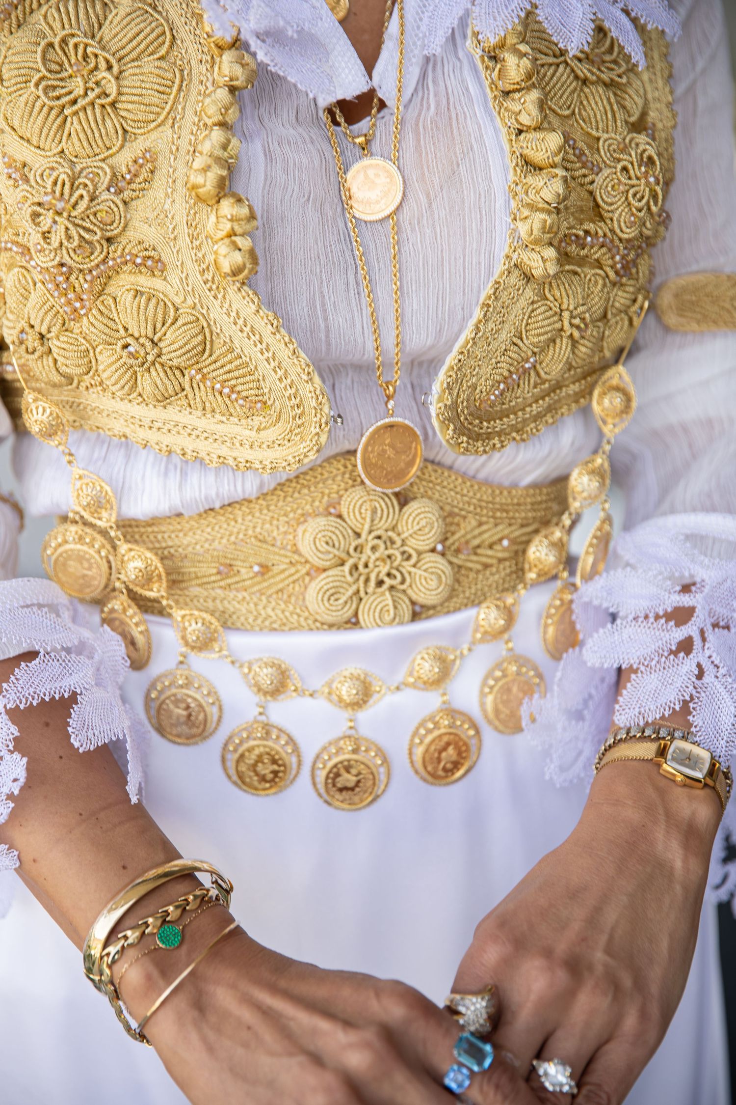 Woman wearing ornate gold embroidered vest with coin jewelry, bracelets, and rings over white lace dress.