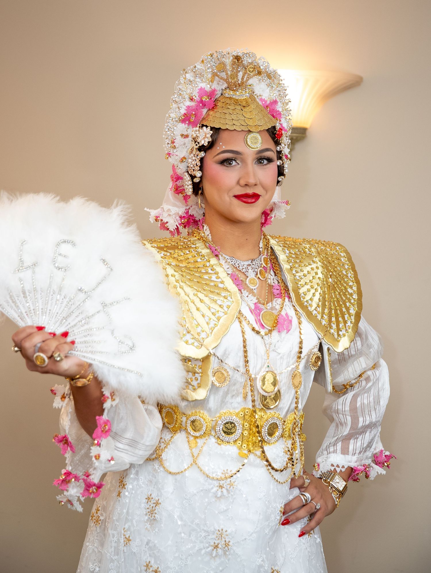 Woman in ornate gold and white traditional costume with floral headdress holding a white feather fan.