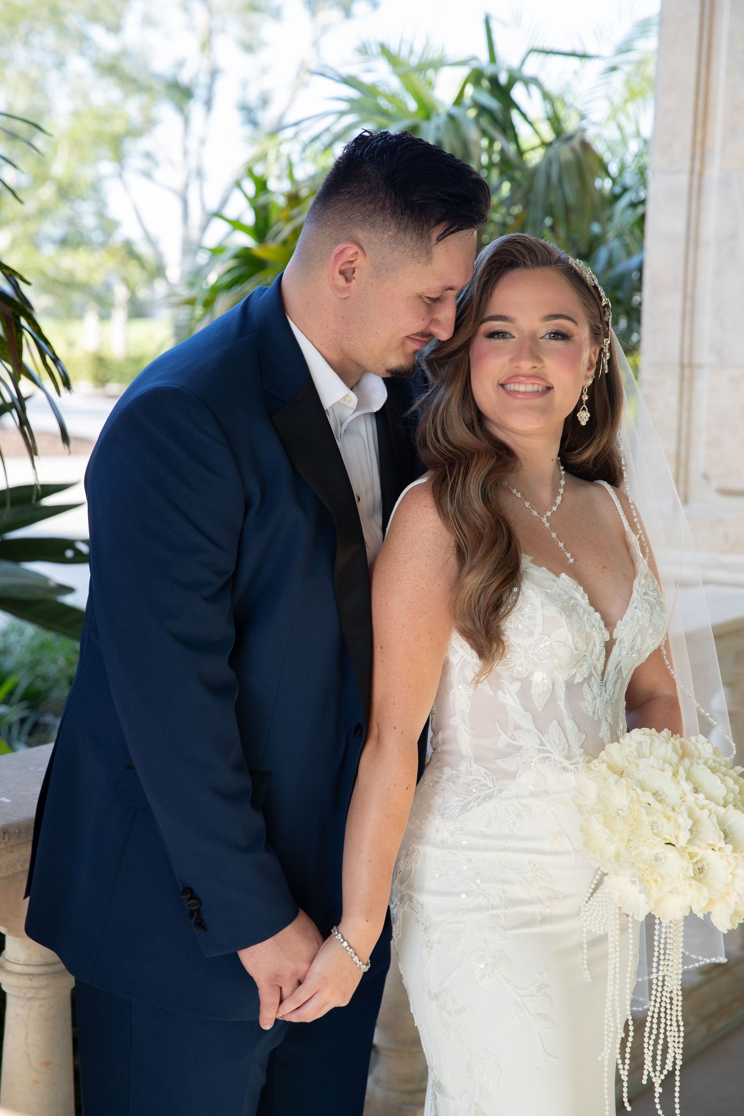 Bride in lace gown holds groom's hand while smiling, holding white bouquet with pearl strands outdoors.