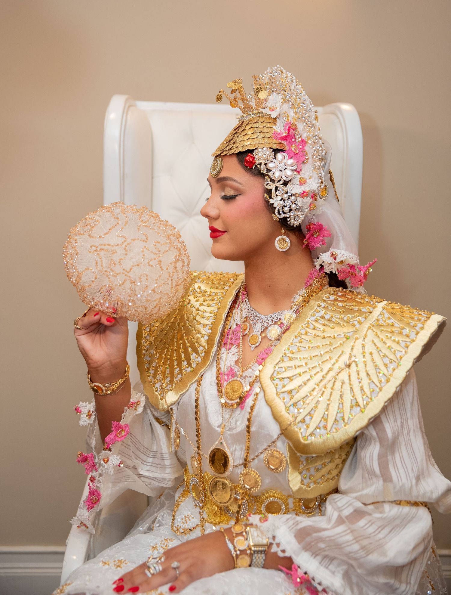 A bride in ornate gold and white traditional attire with floral headpiece holds a decorative fan while seated.