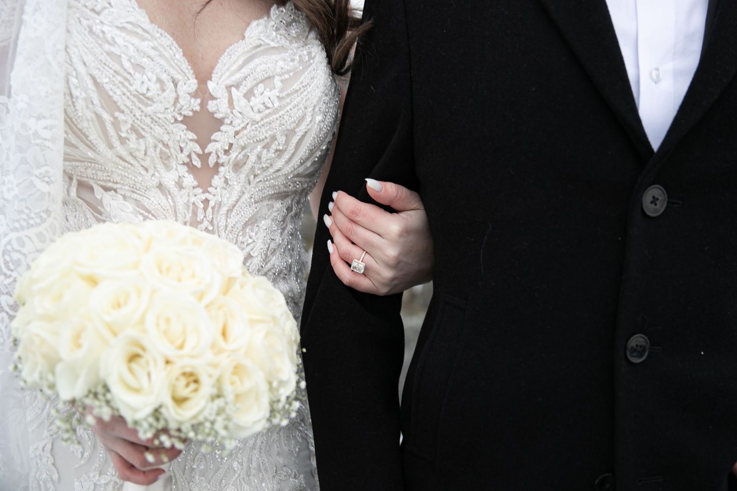 Bride in lace wedding gown holding white rose bouquet stands beside groom in black suit at wedding ceremony.