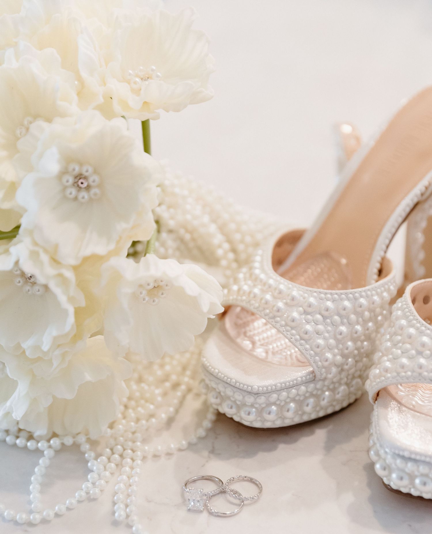 Pearl-covered bridal heels beside white floral bouquet and diamond wedding rings on white surface.