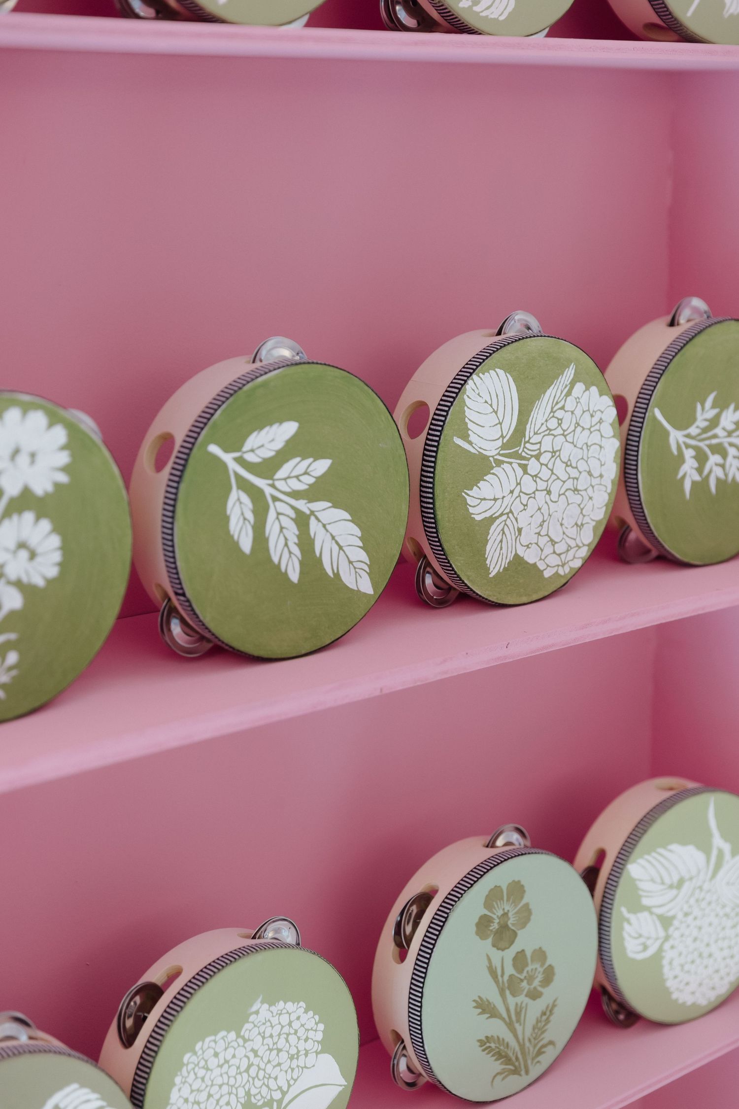 Green and pink floral tambourines displayed on pink shelves, featuring white botanical designs.
