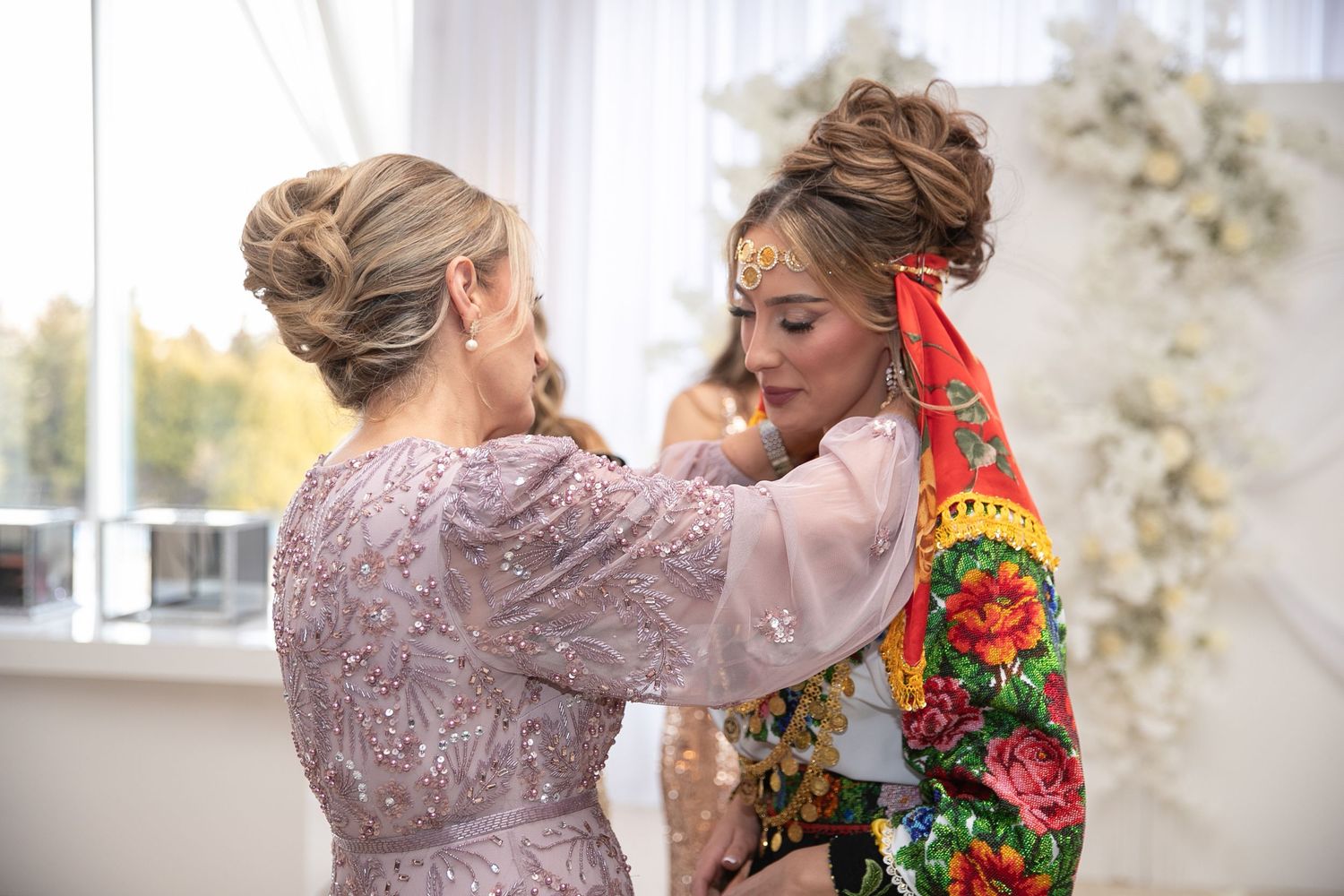 Two women in elegant attire, one in traditional floral folk costume, share a tender moment at a formal celebration.