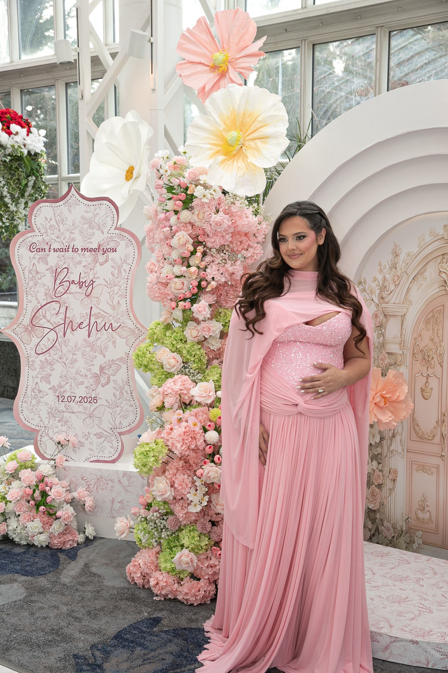 Pregnant woman in pink gown at baby shower for Baby Shelia, surrounded by floral decorations and large paper flowers.