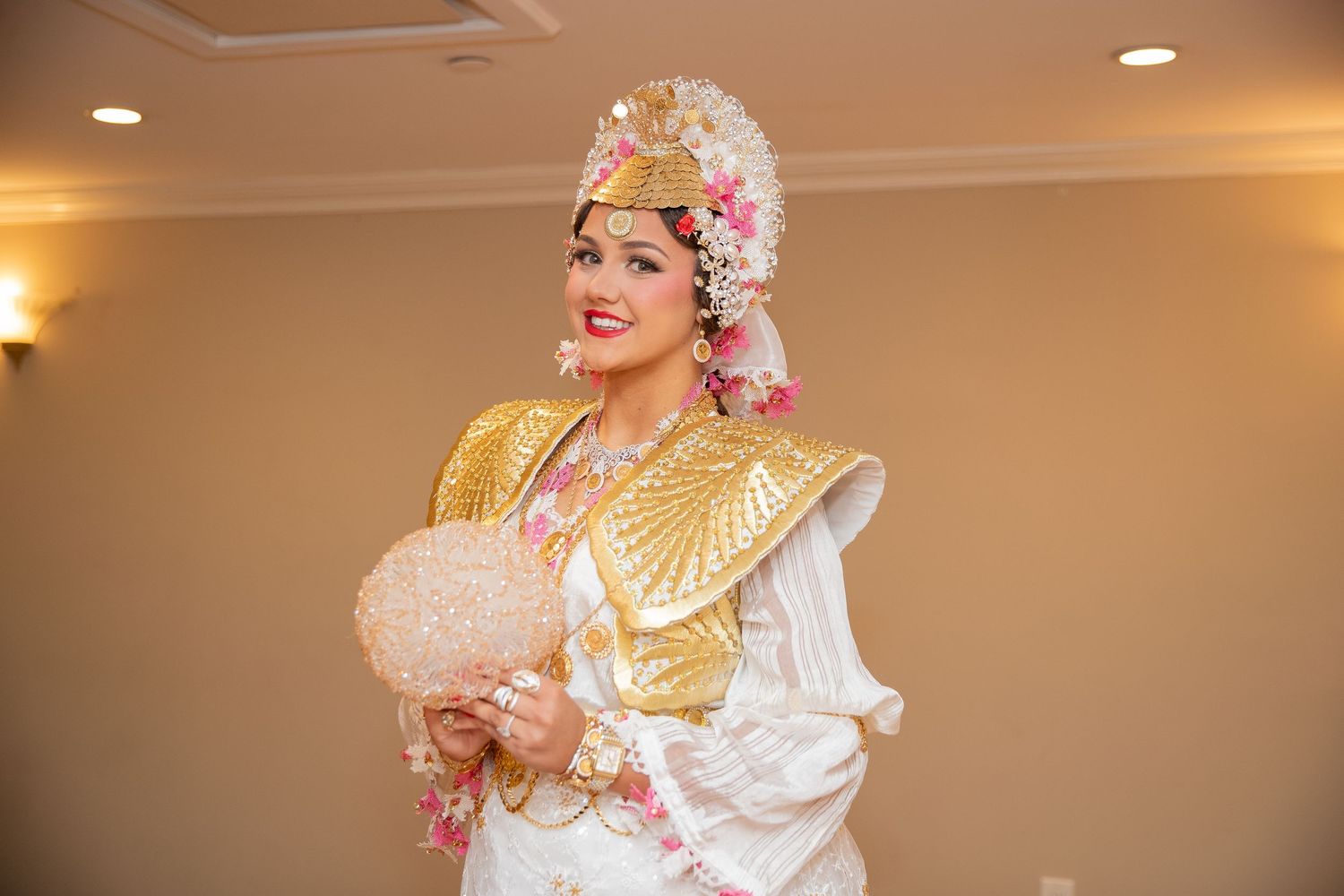 Woman in ornate traditional costume with gold armor, floral headdress, and fan at a cultural event.