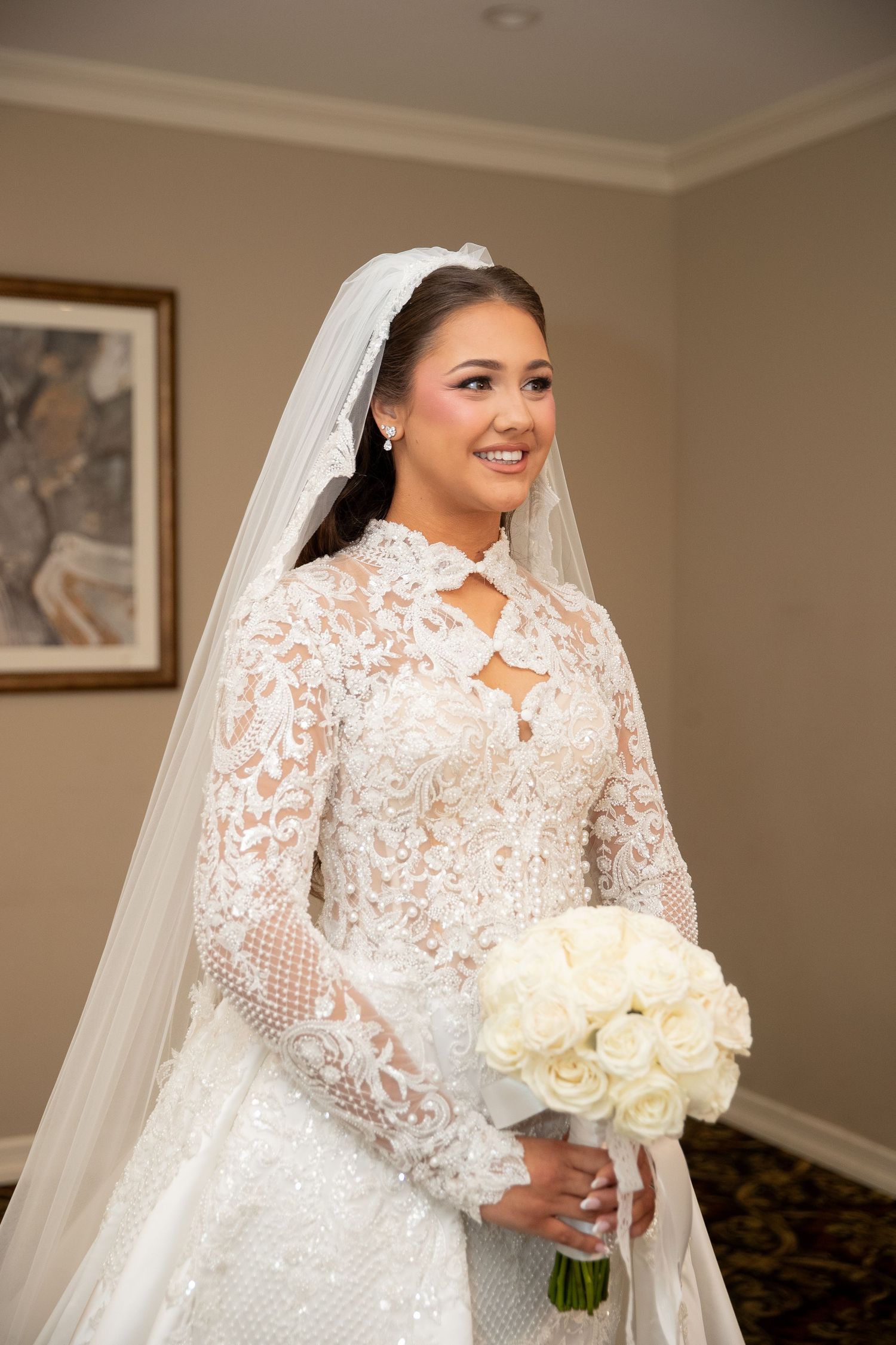 Smiling bride in long-sleeve lace wedding gown and veil holding white rose bouquet indoors.