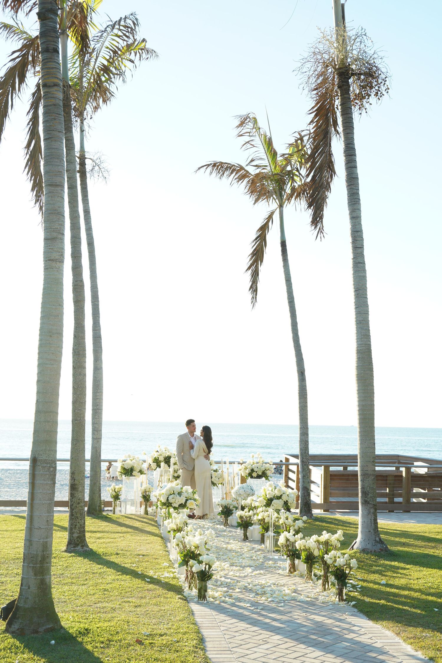 Romantic beach wedding setup with white flowers lining a path between palm trees at sunset.