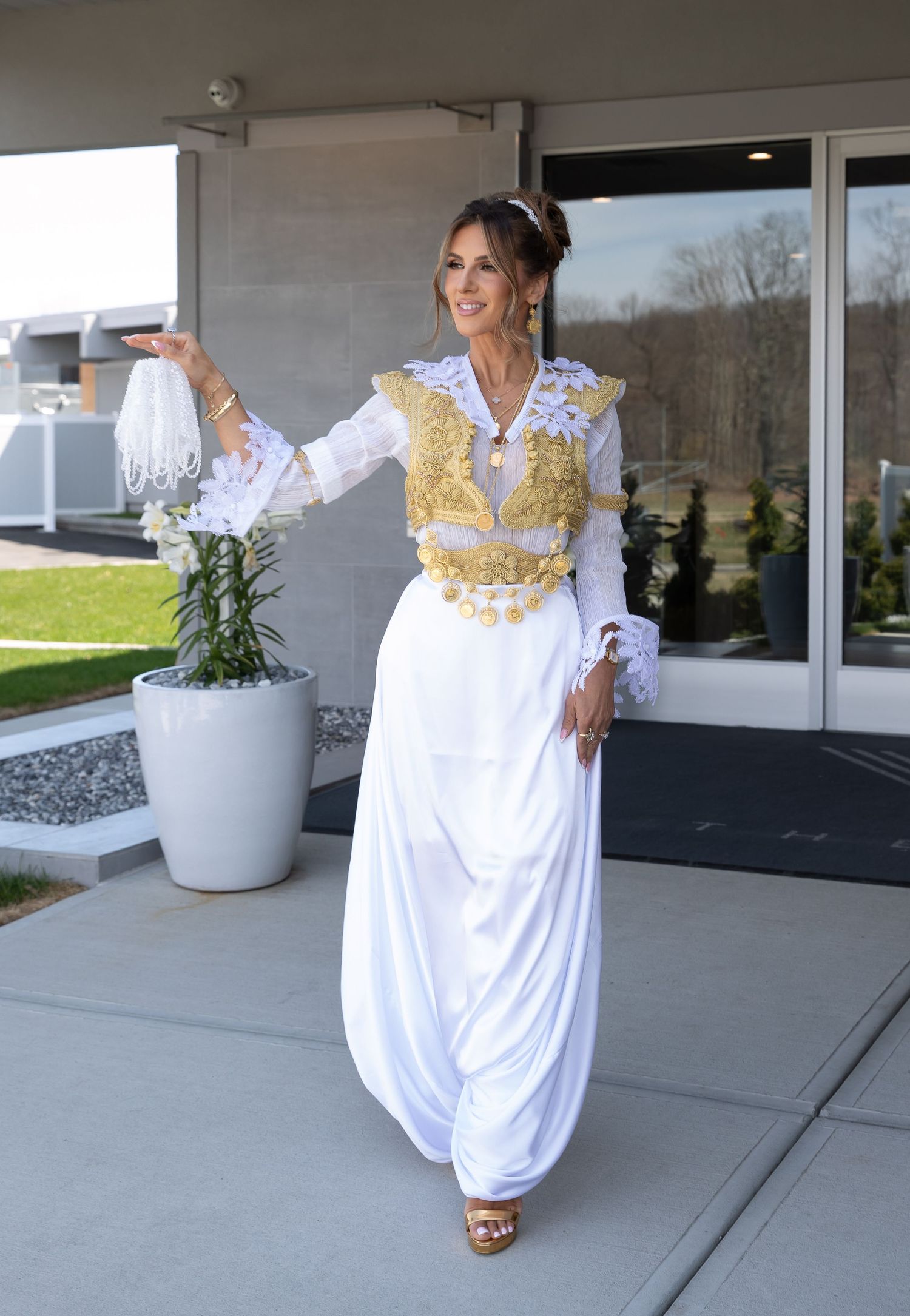Woman in elegant white and gold traditional dress posing gracefully outdoors near a modern building entrance.