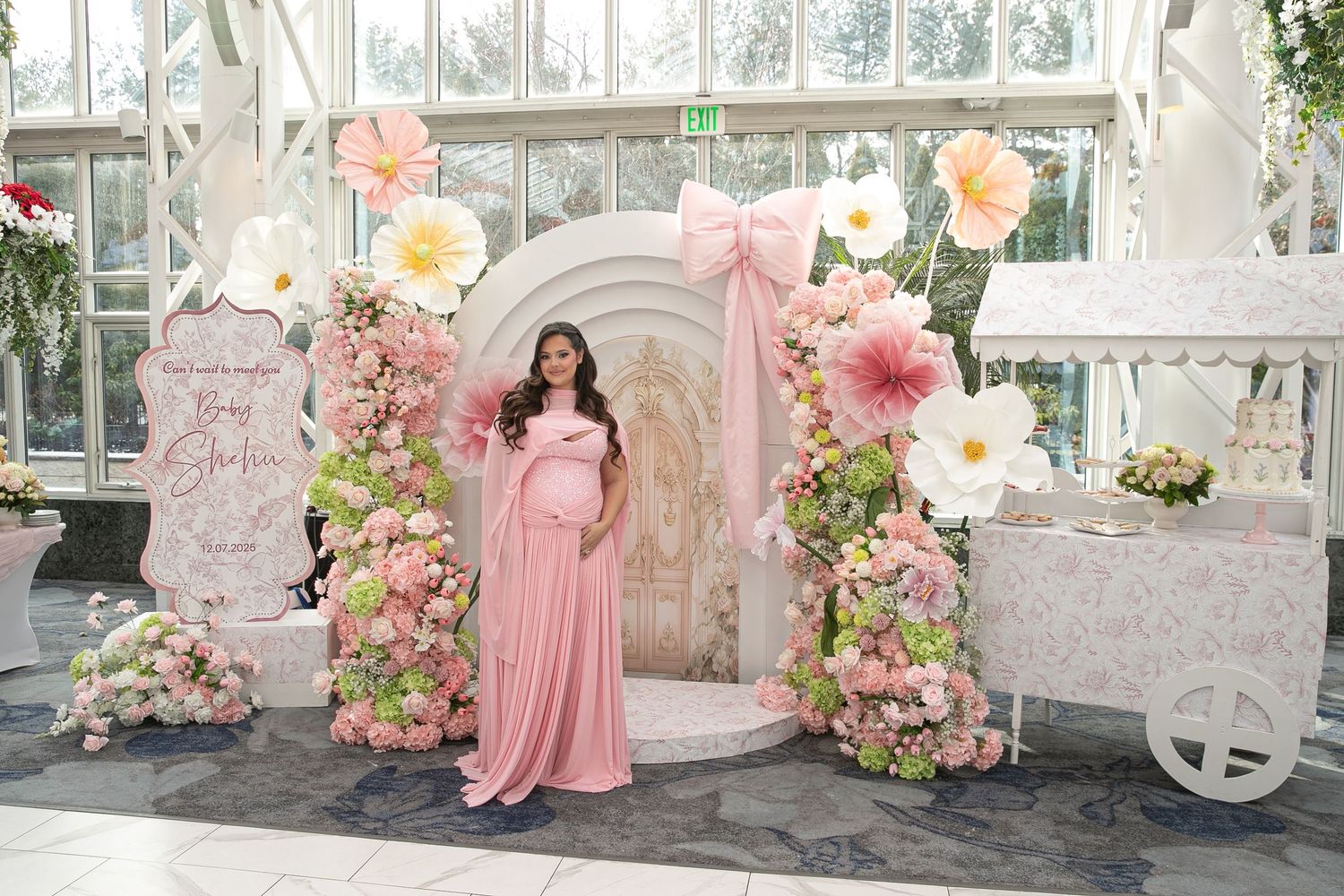 Pregnant woman in pink gown poses at floral baby shower backdrop with large paper flowers and pink bow arch.