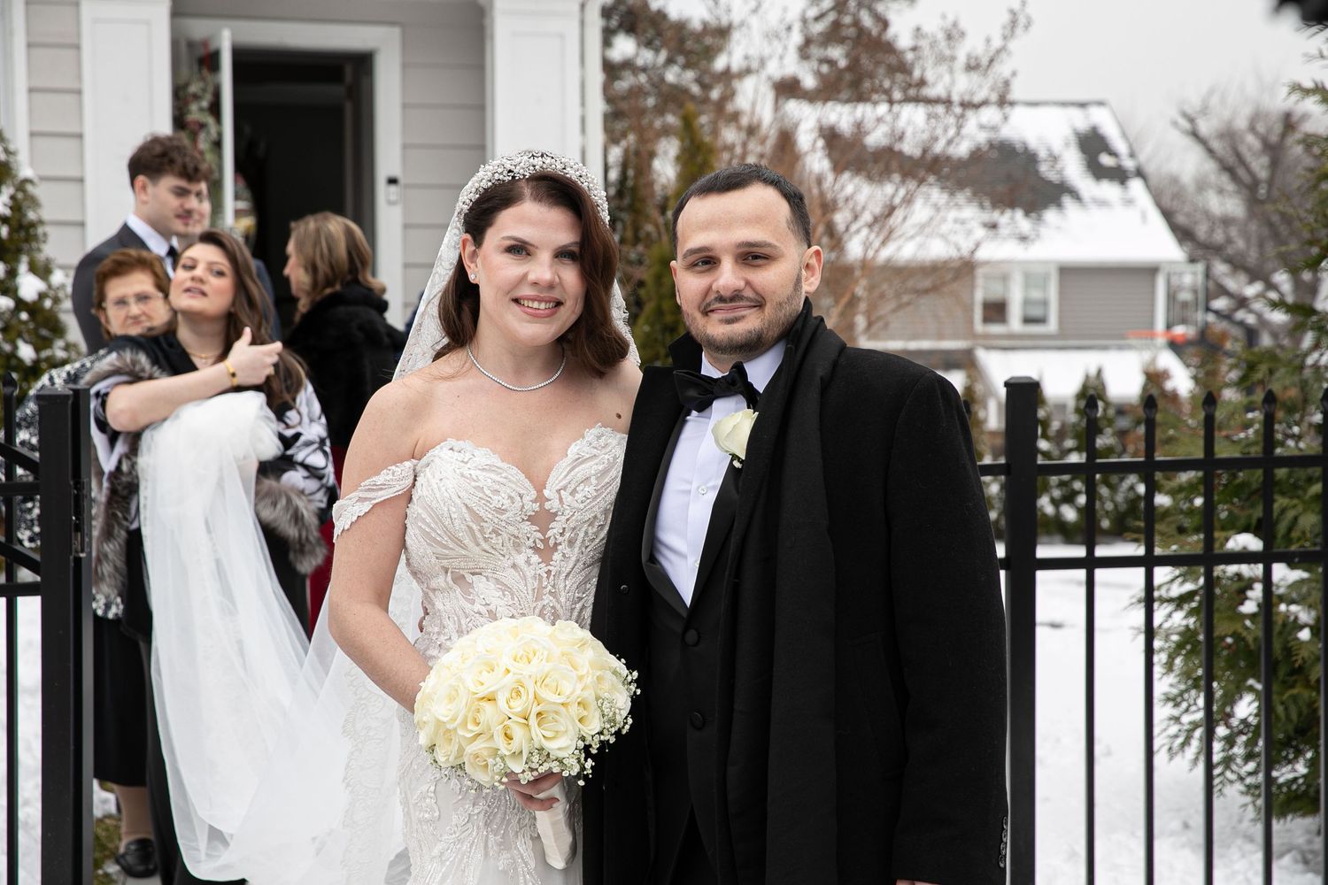 Bride and groom posing outdoors in winter, bride wearing white gown with bouquet, groom in black tuxedo.