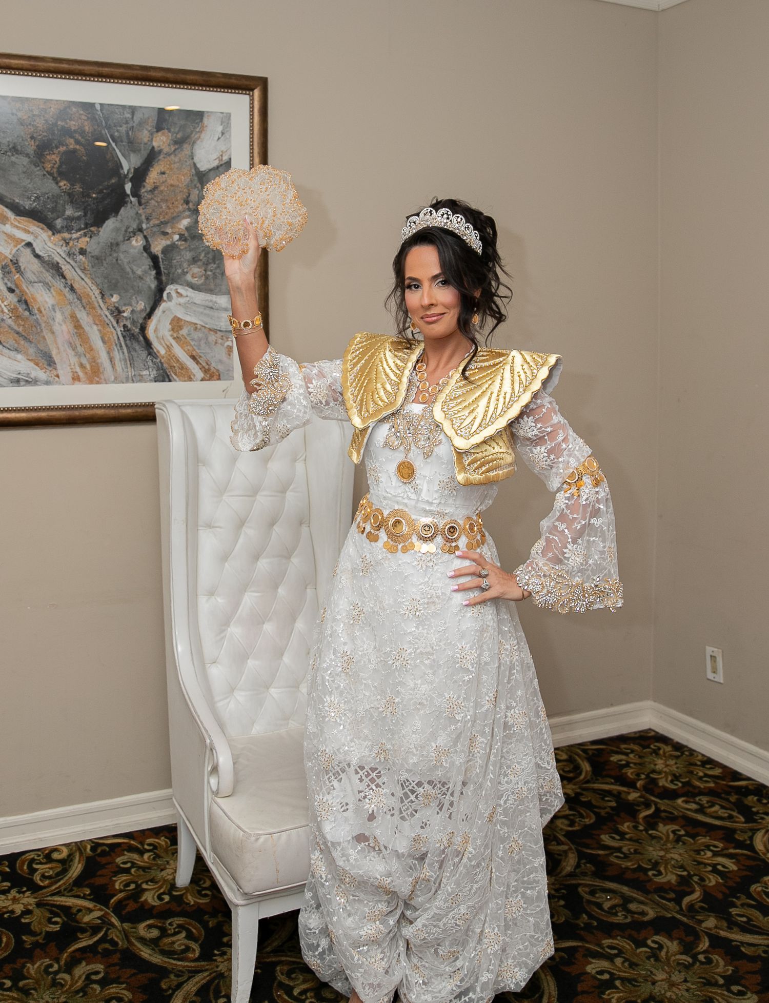 Bride in white lace gown with gold embroidered bolero jacket and tiara holds bouquet beside ornate white chair.