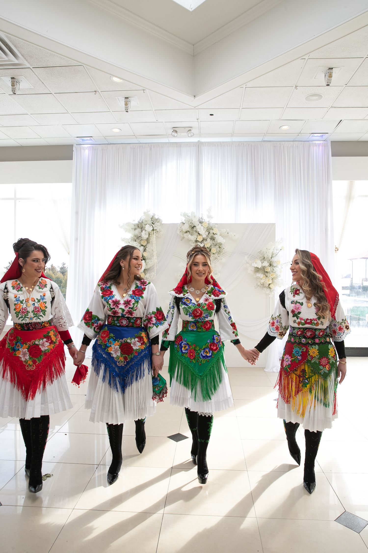 Four women in traditional Eastern European folk costumes with floral embroidery holding hands at a celebration.