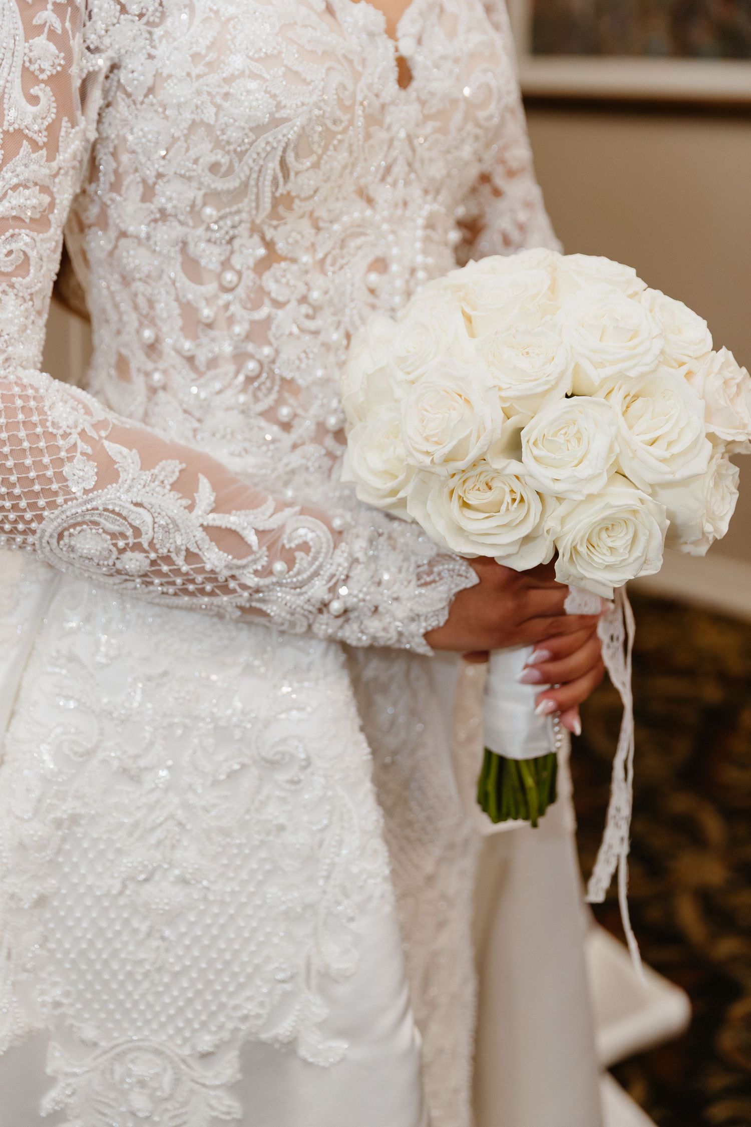 Bride in lace wedding gown holding elegant white rose bouquet tied with ribbon.