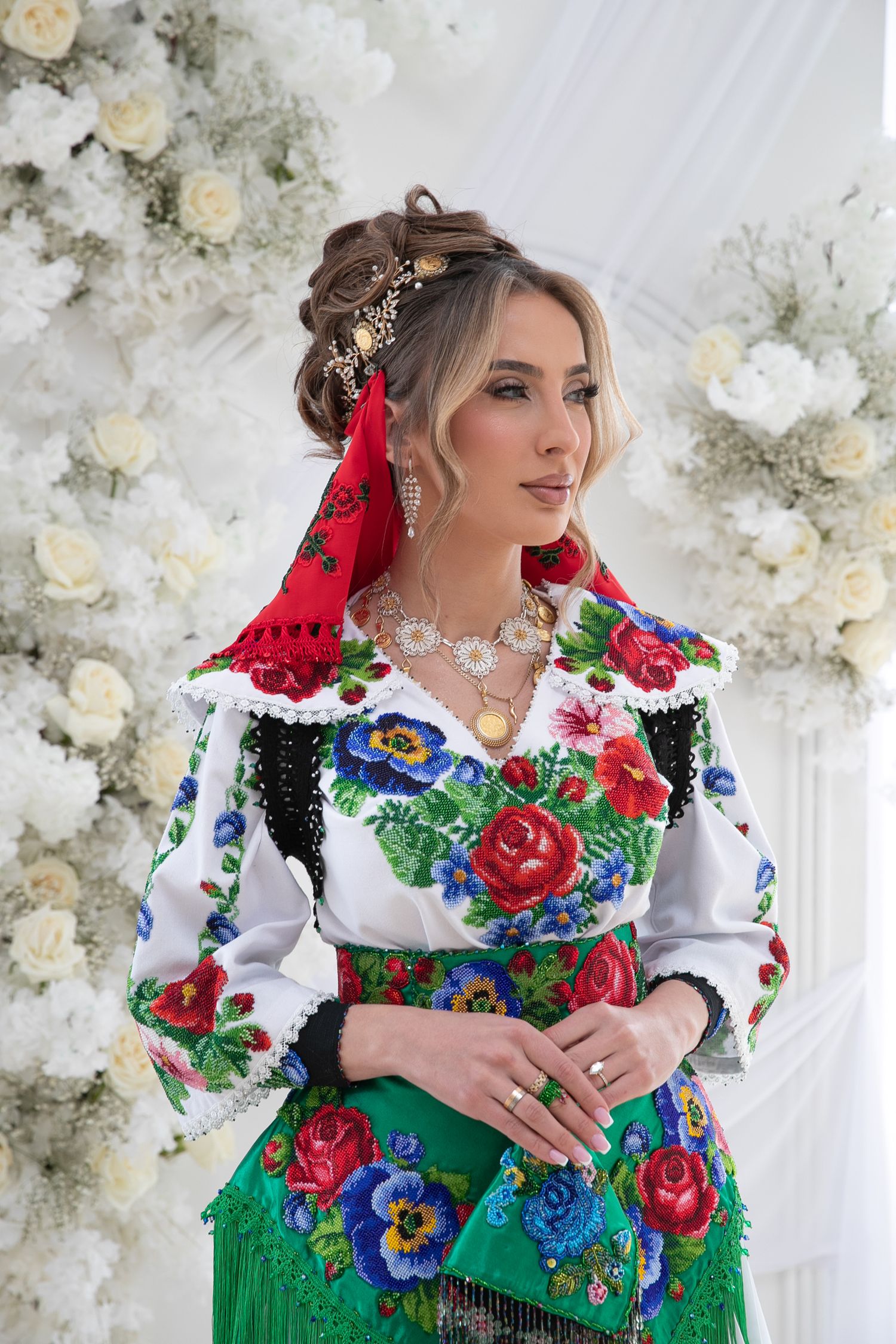 Woman in colorful traditional Eastern European folk costume with floral embroidery and red headscarf, posing near white flowers.