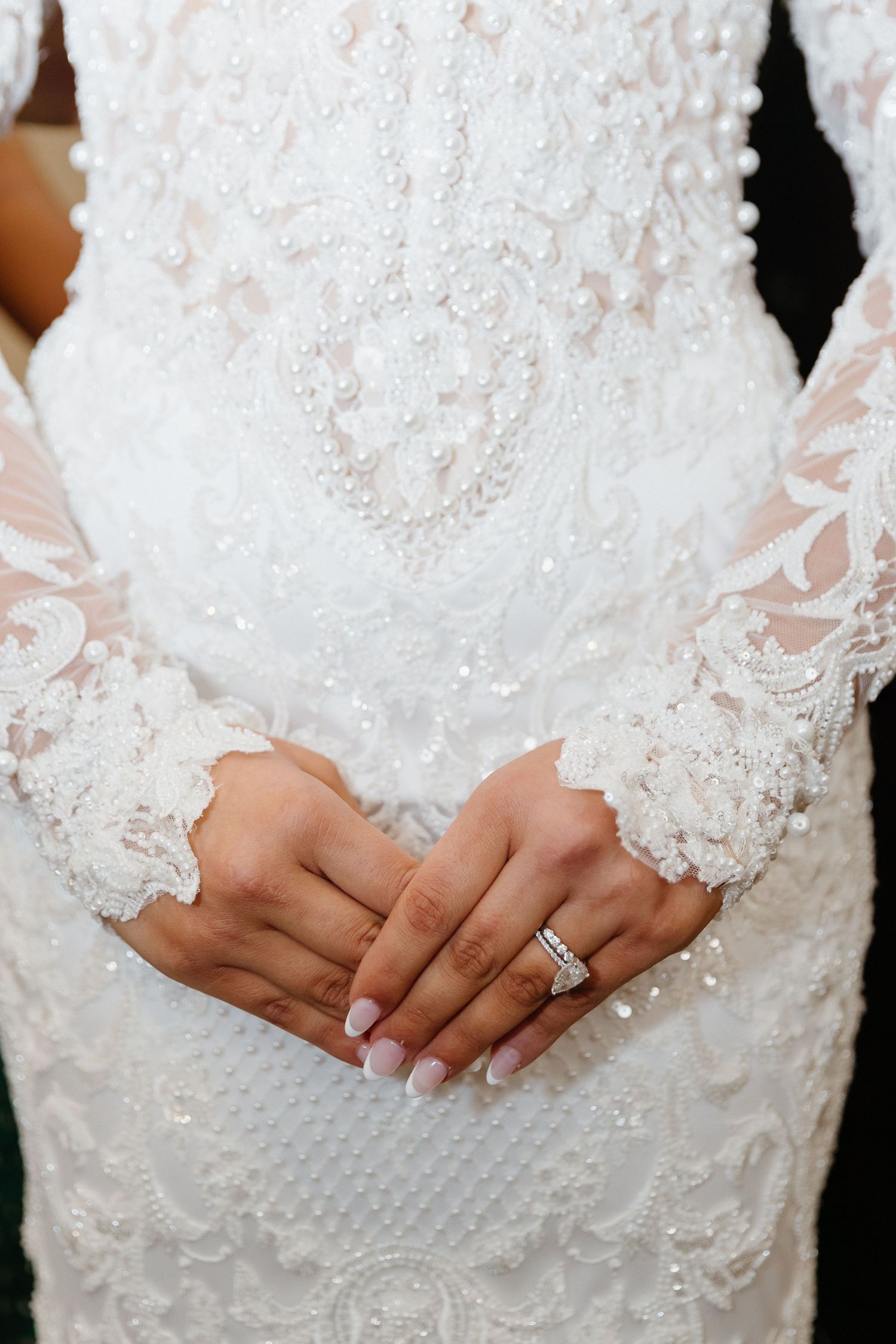 Bride in beaded lace wedding gown with long sleeves, hands clasped showing diamond wedding ring.
