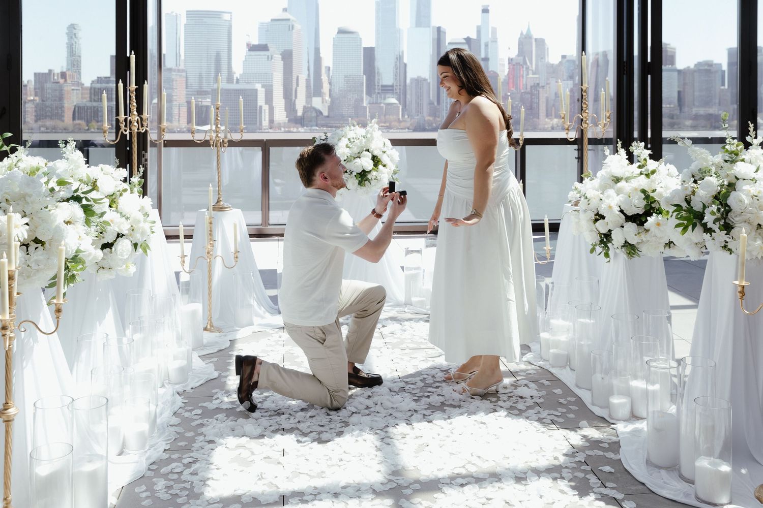 Man proposes on one knee in elegant white floral venue with NYC skyline backdrop.