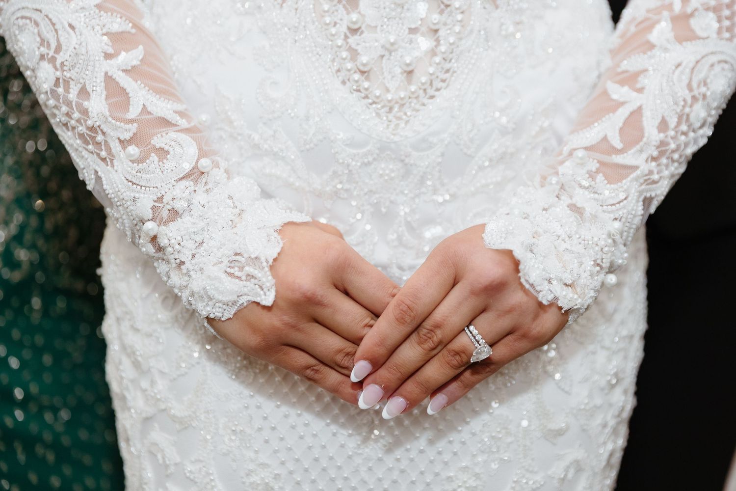 Bride in white lace wedding dress with clasped hands showing diamond engagement ring and manicured nails.