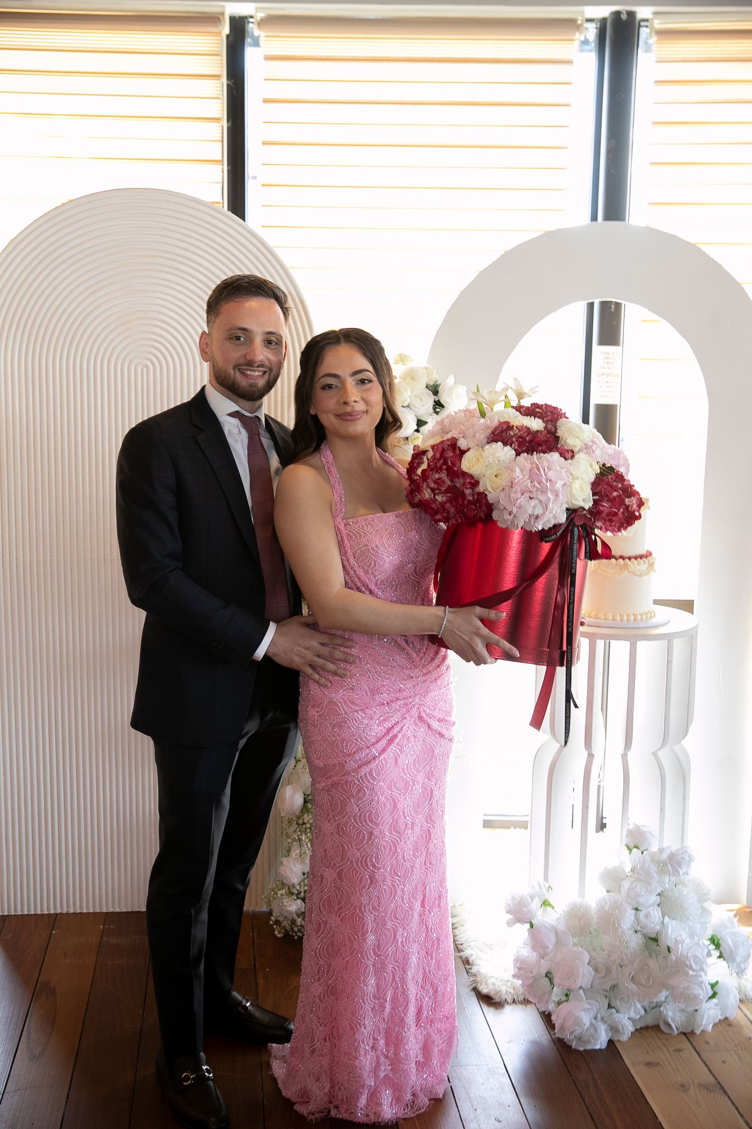 Couple in formal attire posing with floral bouquet and wedding cake display against white arch backdrop.