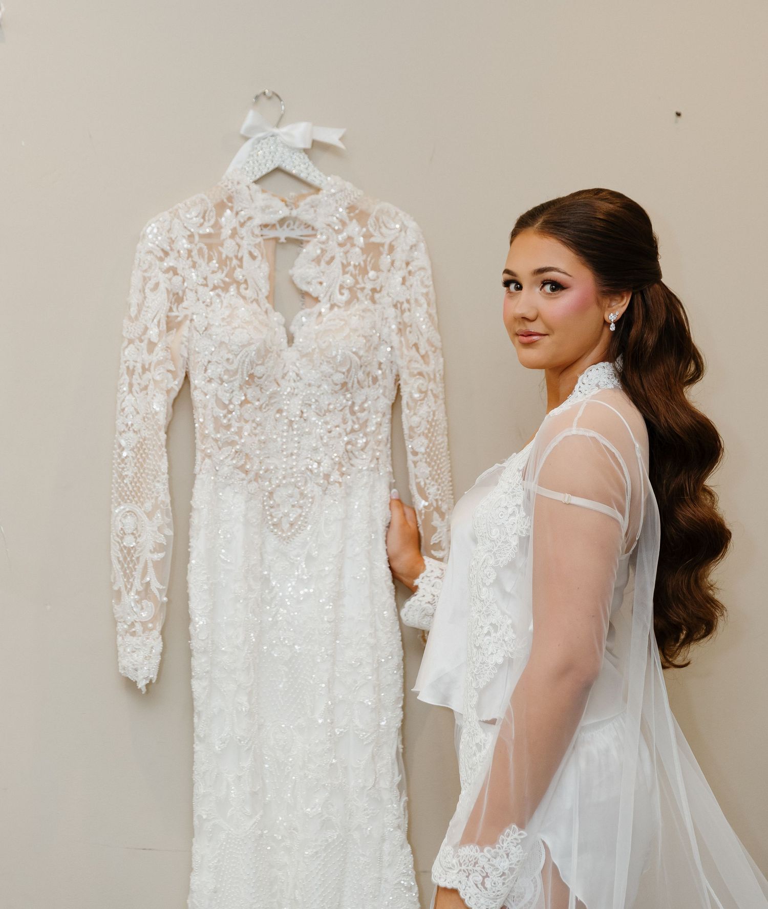 Bride in sheer lace bridal outfit poses beside an elegant long-sleeve lace wedding gown on a hanger.