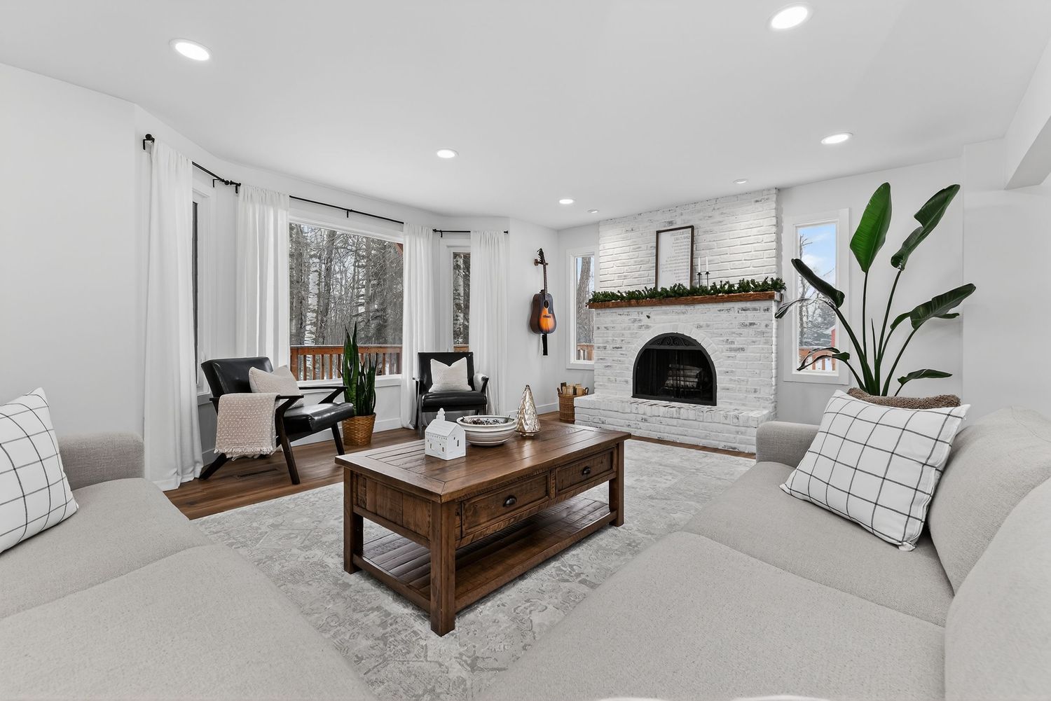 Bright living room with white sofas, brick fireplace, wood coffee table, and large windows with natural light.
