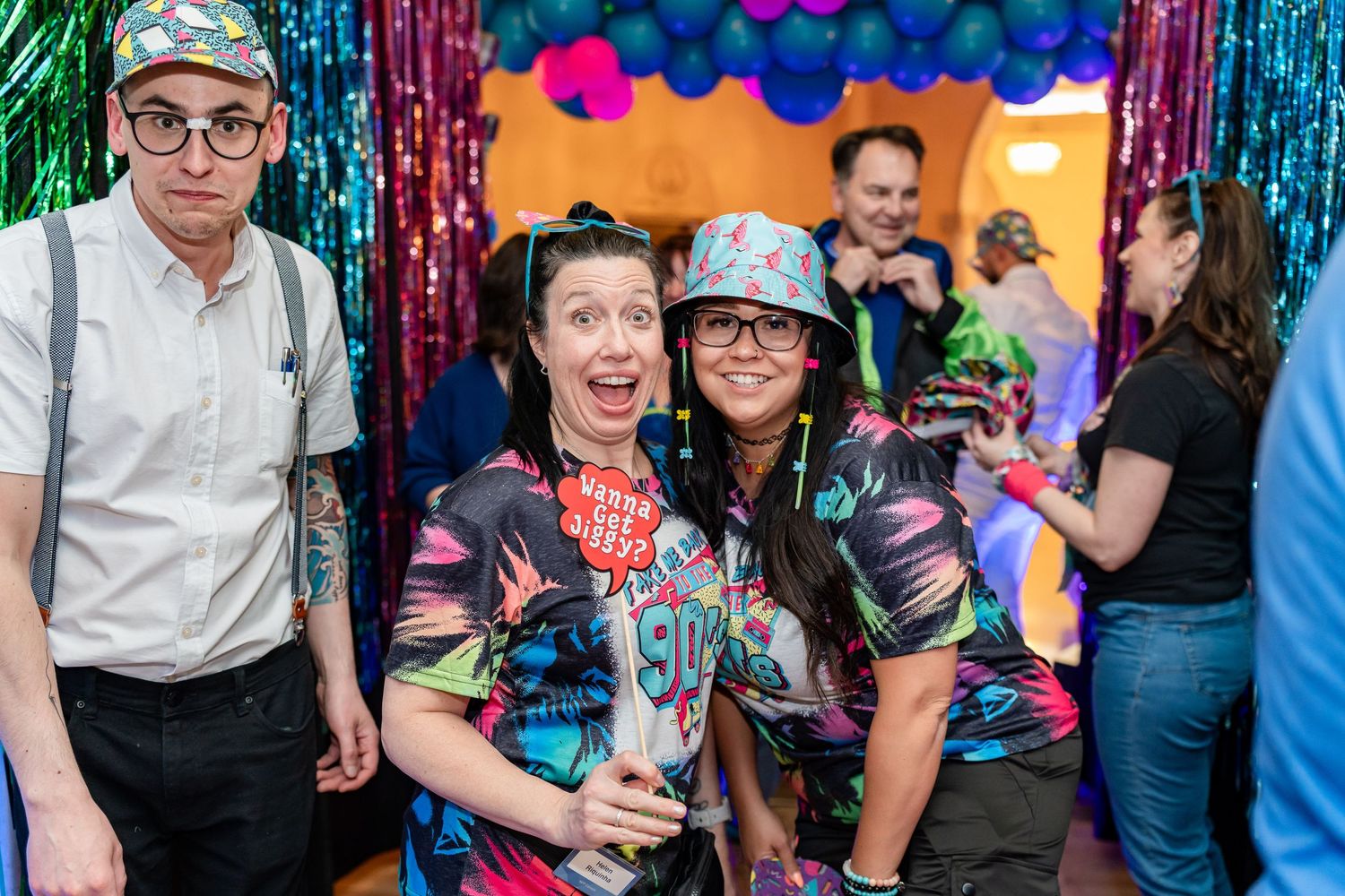 People celebrate at a colorful party photo booth with metallic streamers and balloon decorations.