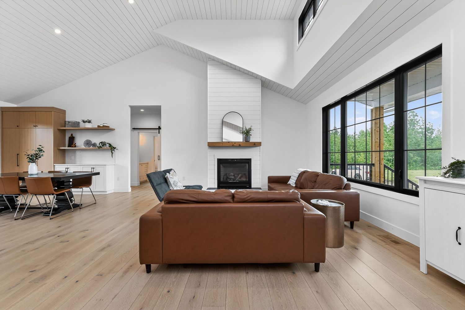 Modern open-concept living room with vaulted ceiling, brown leather sofas, shiplap fireplace, and light hardwood floors.