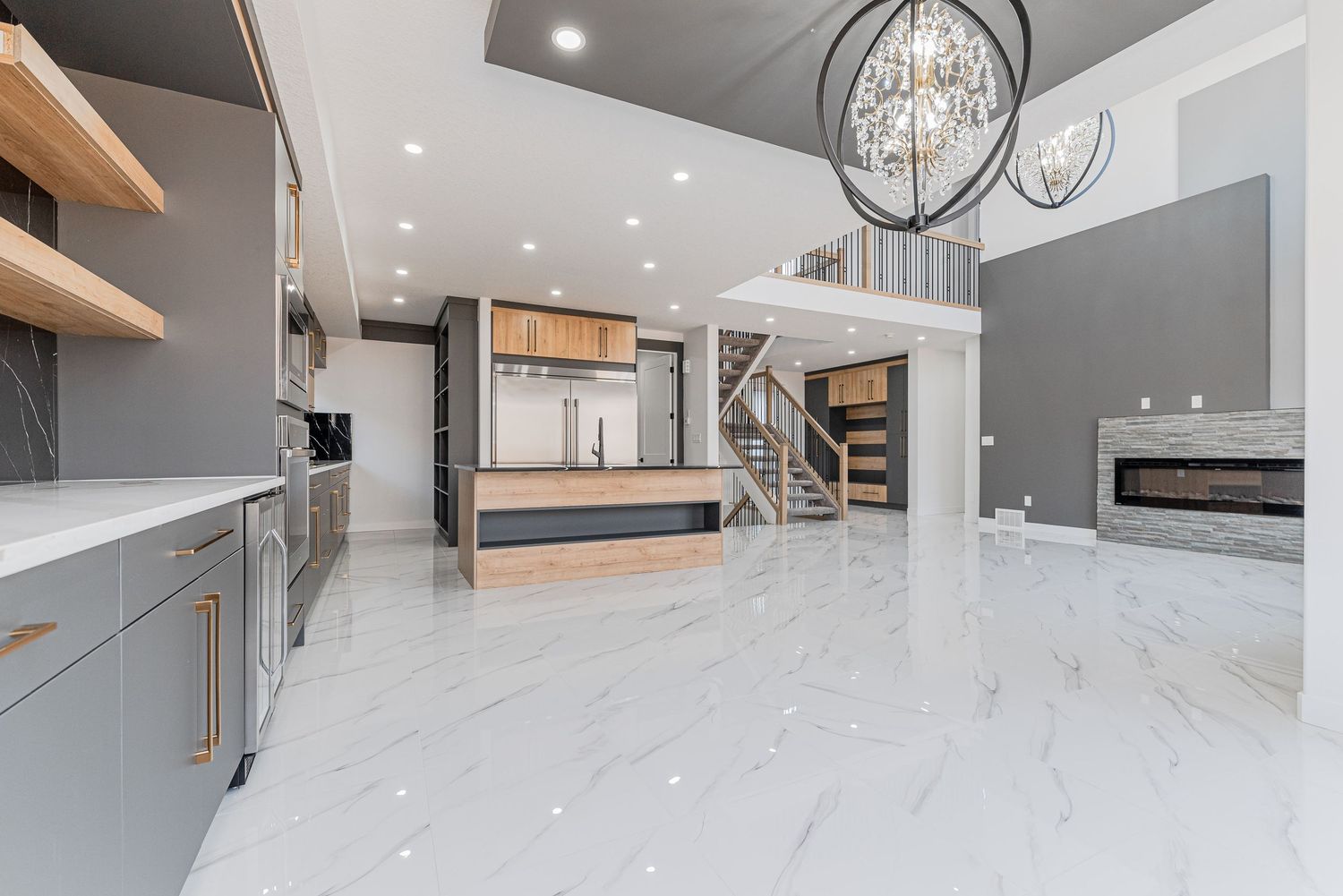 Modern open-concept kitchen with marble floors, gray cabinets, wood accents, chandelier, and fireplace in luxury home.