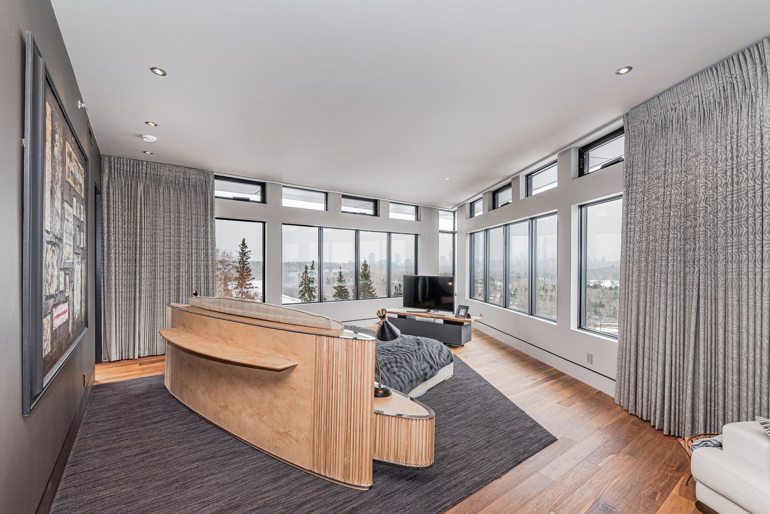 Modern living room with panoramic windows, hardwood floors, curved wood desk, and city views bathed in natural light.