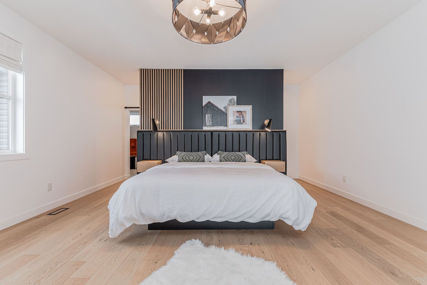 Modern bedroom with white bedding, dark accent wall, light hardwood floors, and a decorative chandelier.