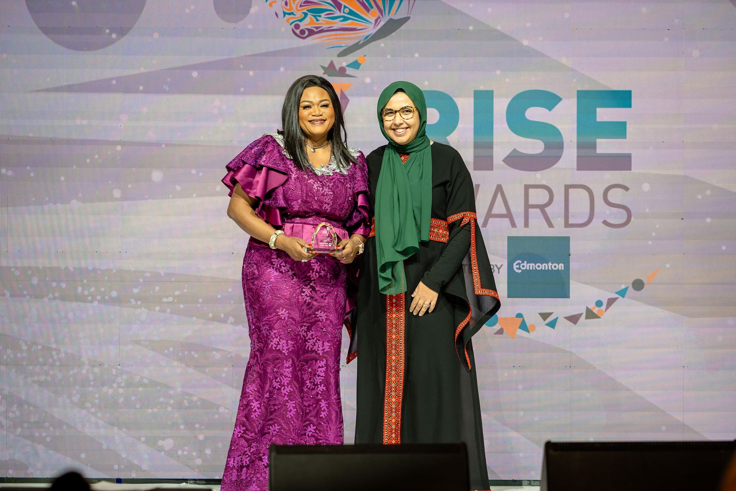 Presenters on stage at the RISE Awards wearing formal attire against purple backdrop.