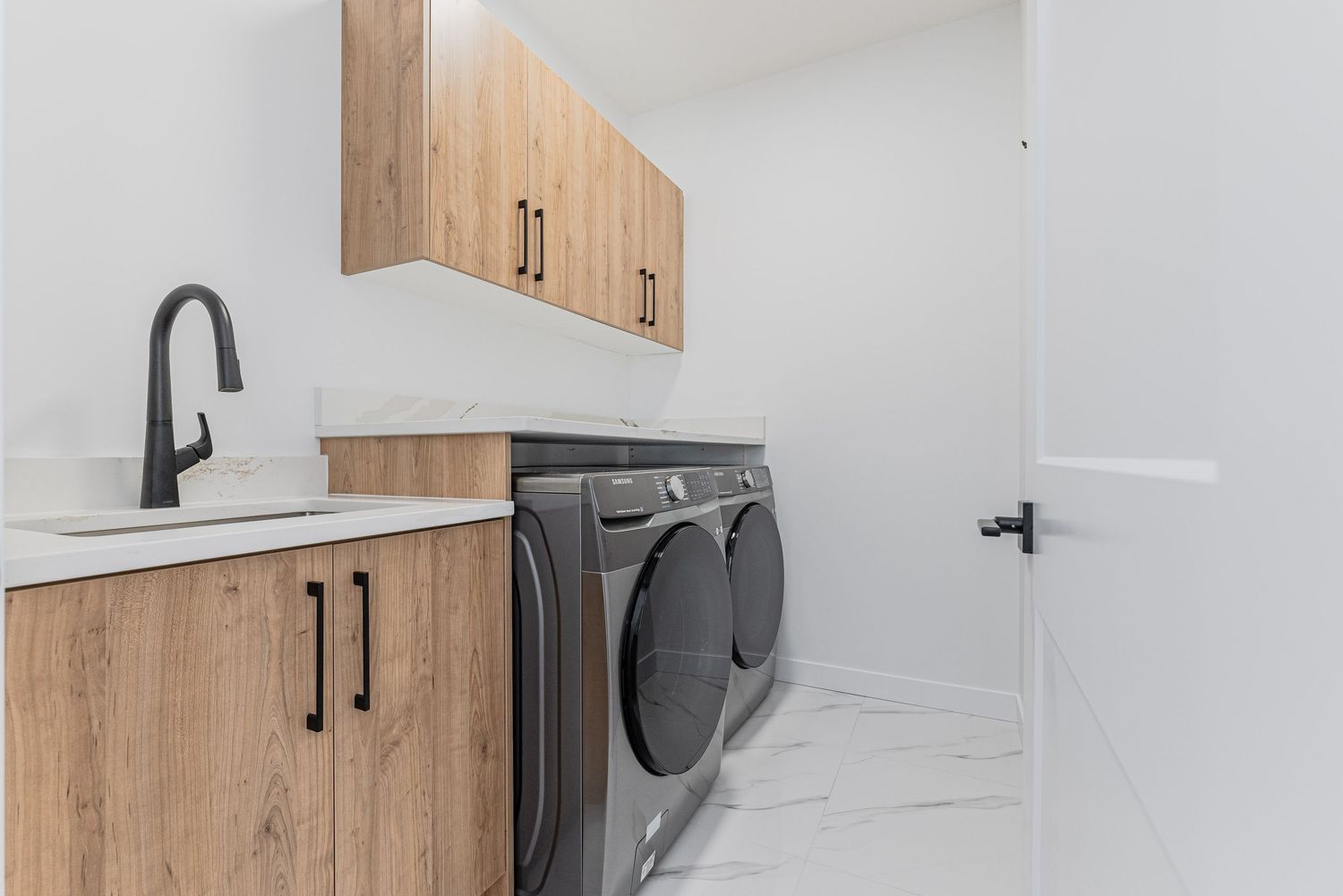 Modern laundry room with wood cabinets, front-load washer and dryer, marble tile floors, and a utility sink with black faucet.