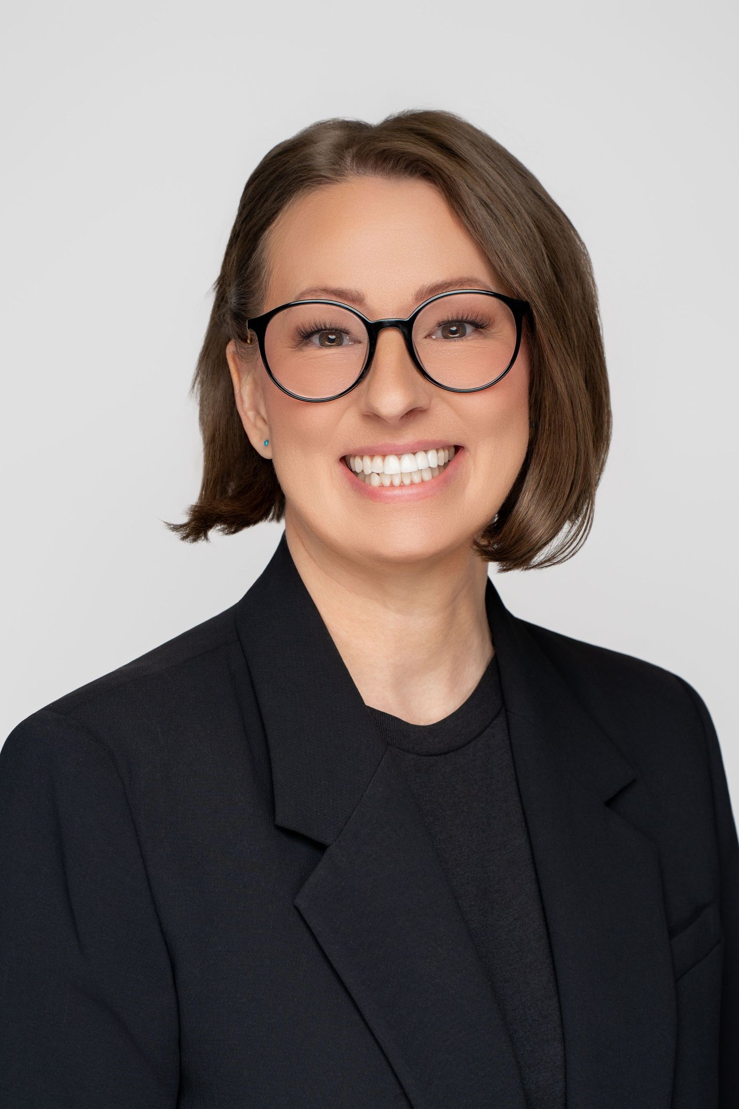 Professional headshot with short brown hair wearing glasses and black blazer with friendly expression.