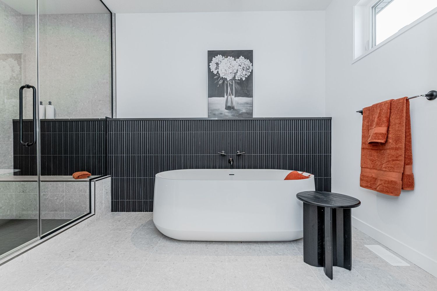 Modern bathroom with freestanding white tub, dark tile accent wall, orange towels, and glass shower enclosure.