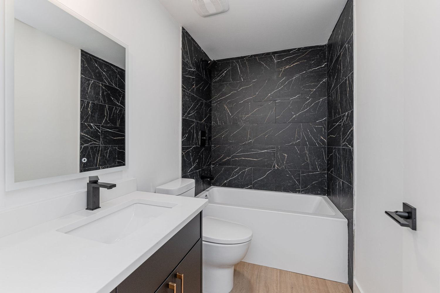 Modern bathroom with black marble tile surround bathtub, white vanity, dark fixtures, and wood-look flooring.