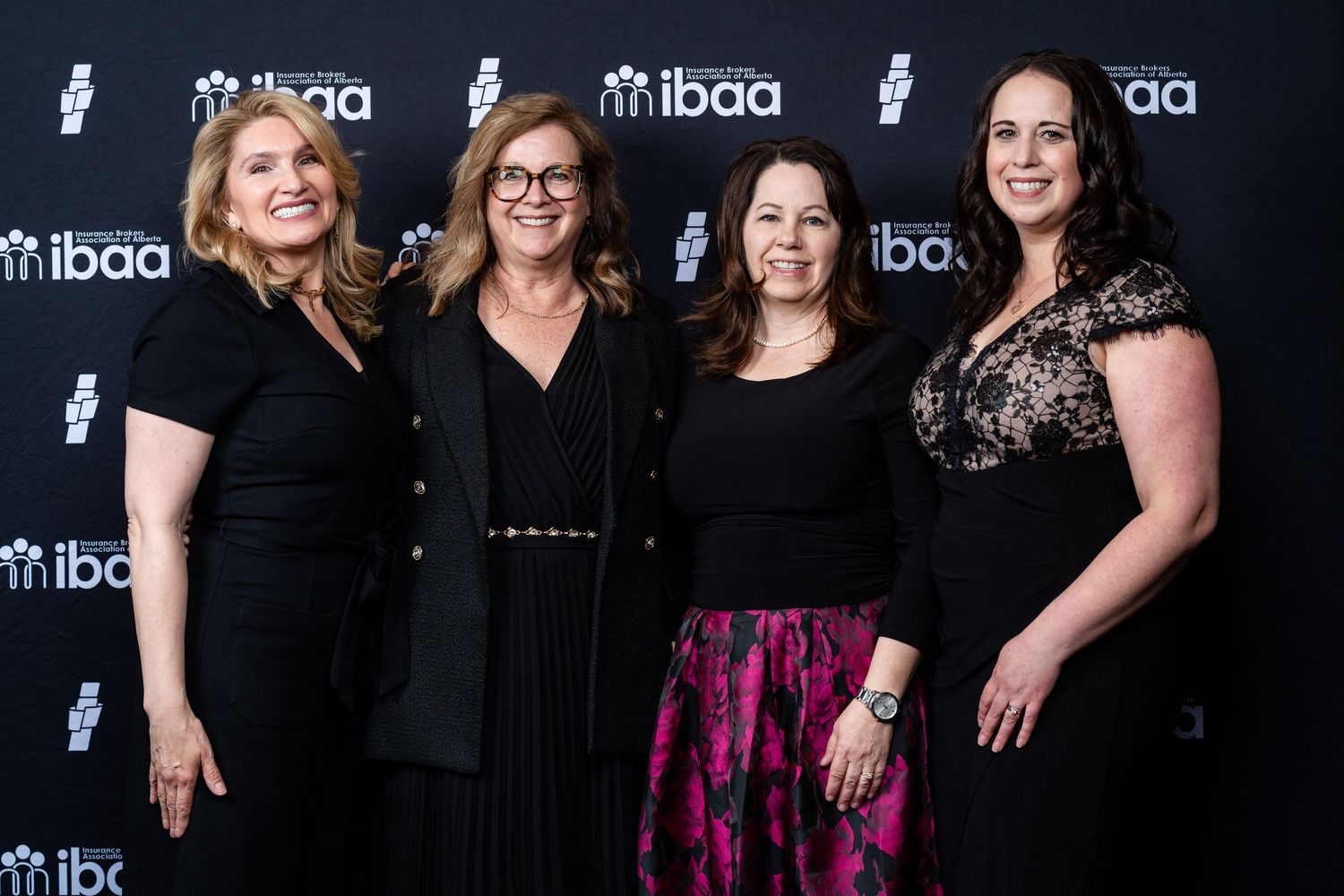 Four women in formal black attire pose together at an IBAA event against a branded backdrop.