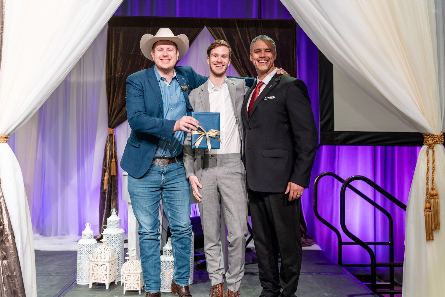 Three people stand under white drapes at an awards ceremony, with one wearing a cowboy hat and holding a trophy.