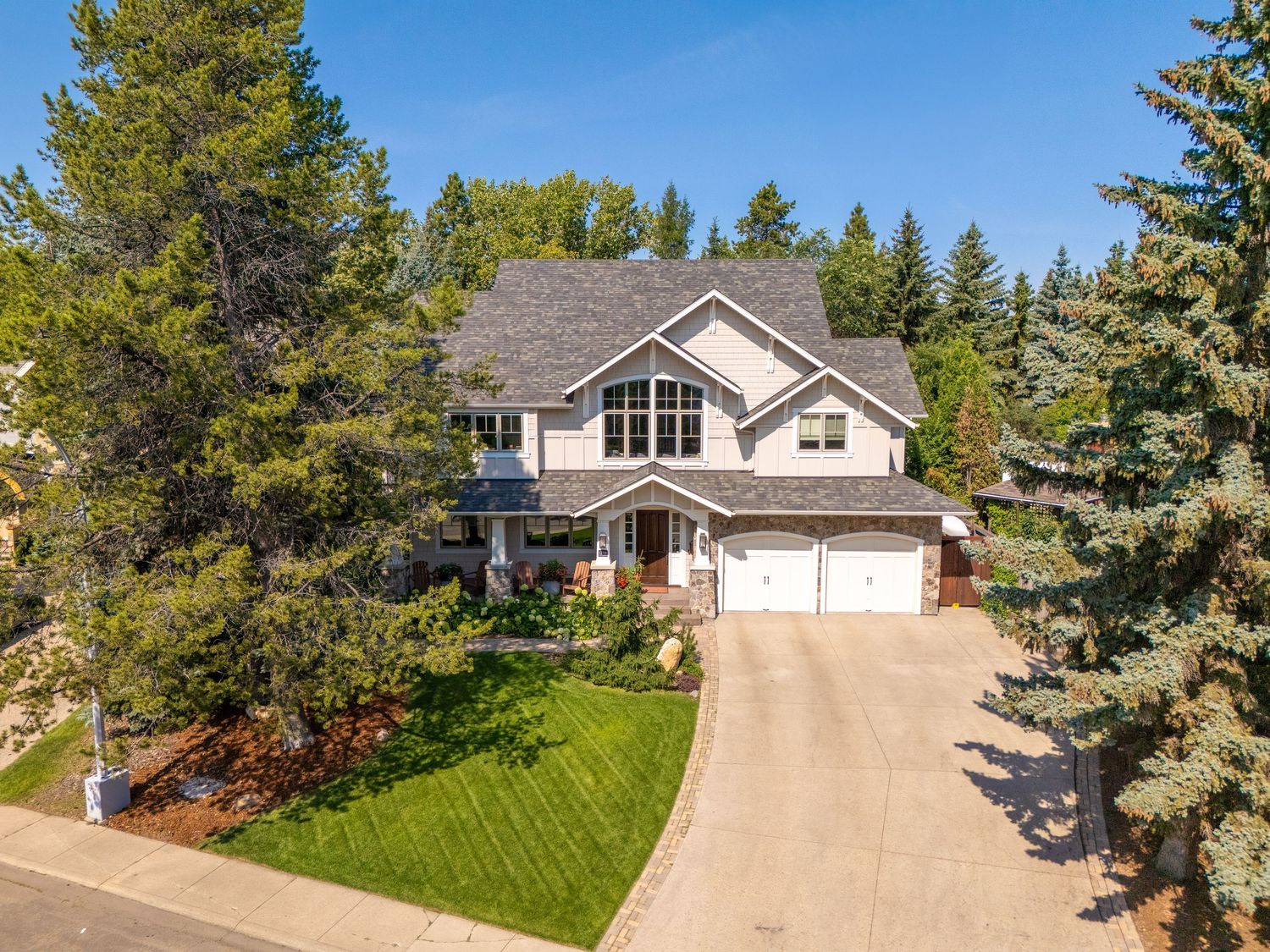Large two-story home with attached double garage surrounded by mature evergreen trees on a curved driveway.