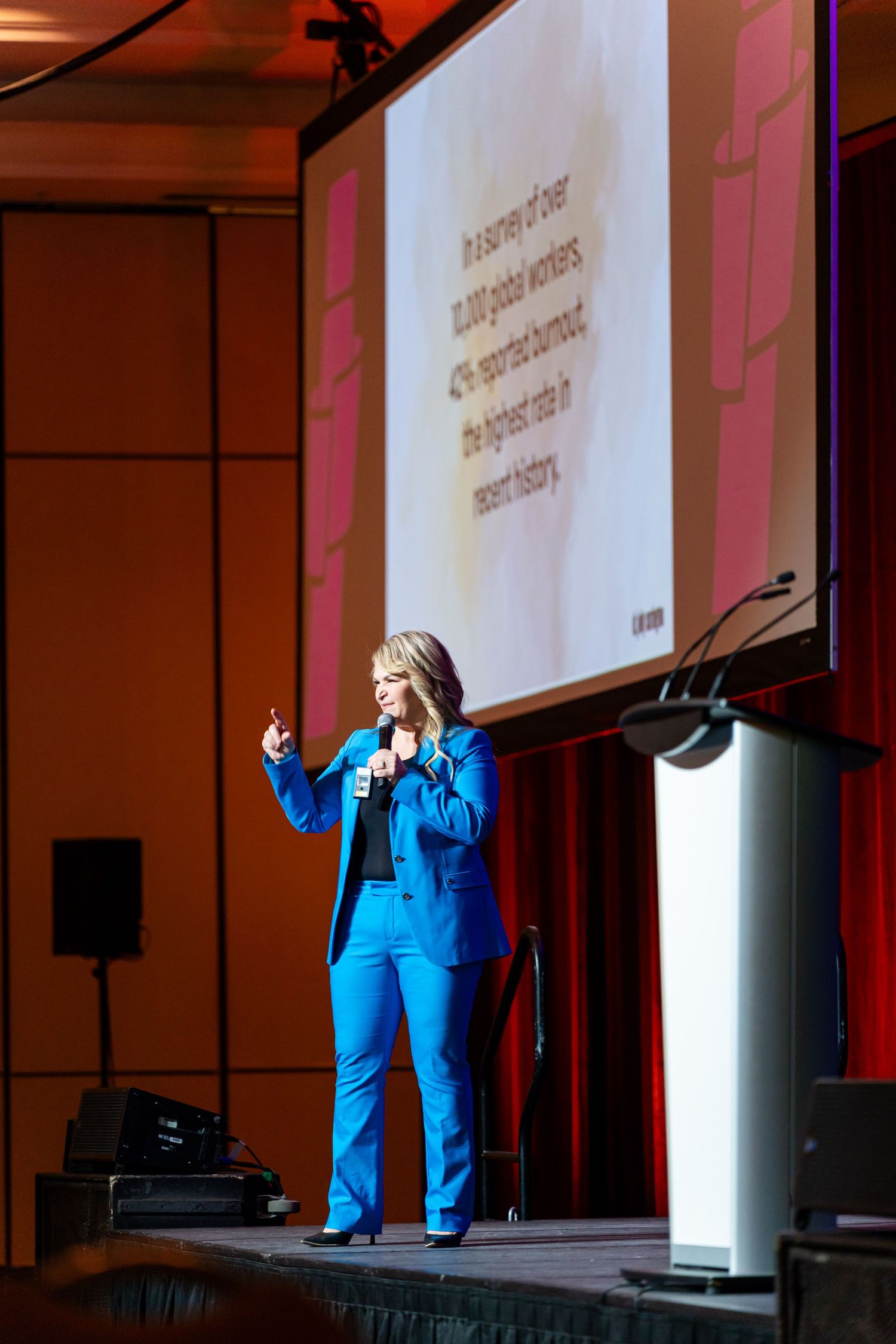Speaker in bright blue attire presenting on stage with projection screen visible.